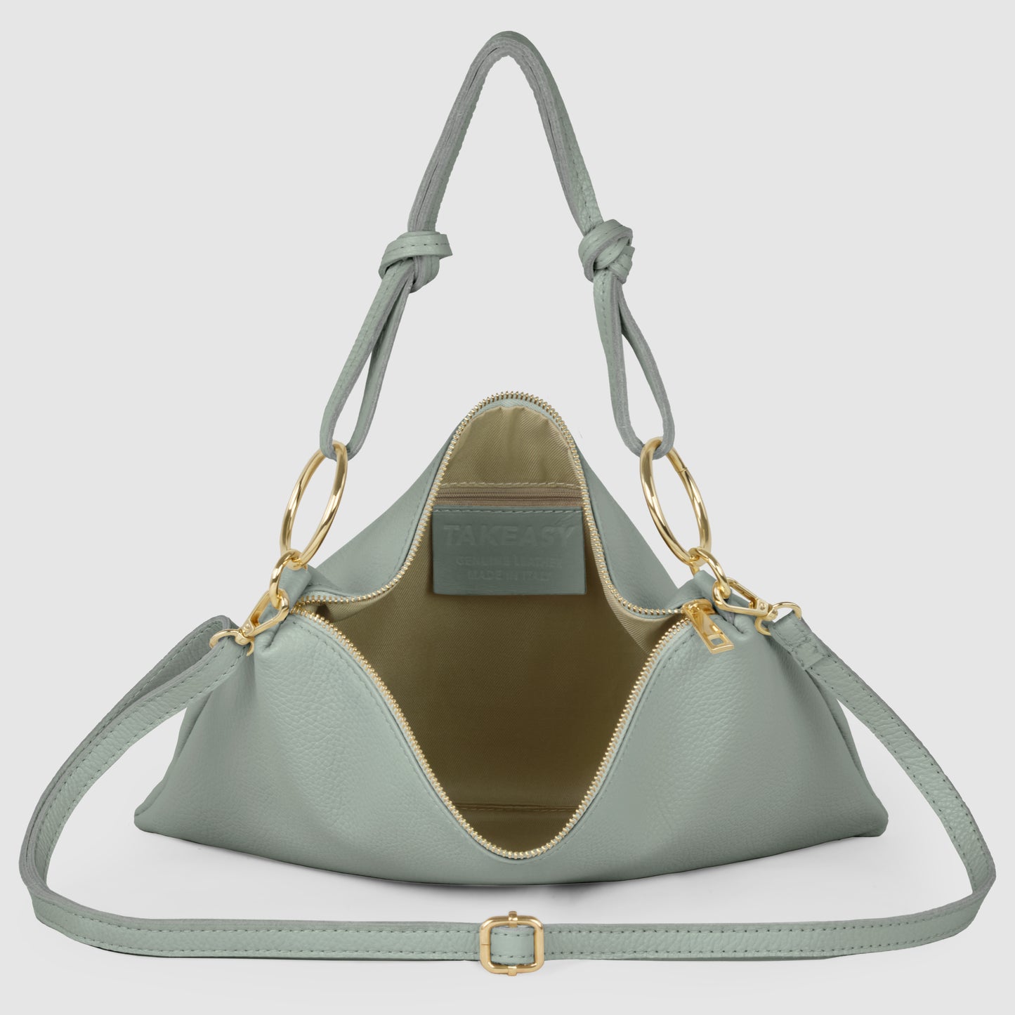 A light sage green Italian leather handbag with gold hardware, an open zipper showing a light brown interior, adjustable shoulder strap, and two knotted handles. The brand label is visible inside this structured zippered purse.