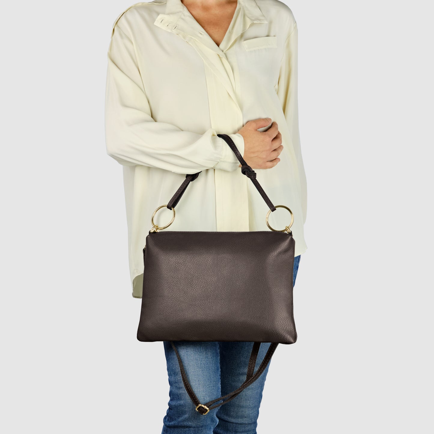 A person wearing a cream-colored blouse and blue jeans holds an elegant crossbody bag with gold rings and a shoulder strap. The structured zippered handbag, in black leather, hangs at waist level against a plain, light background.