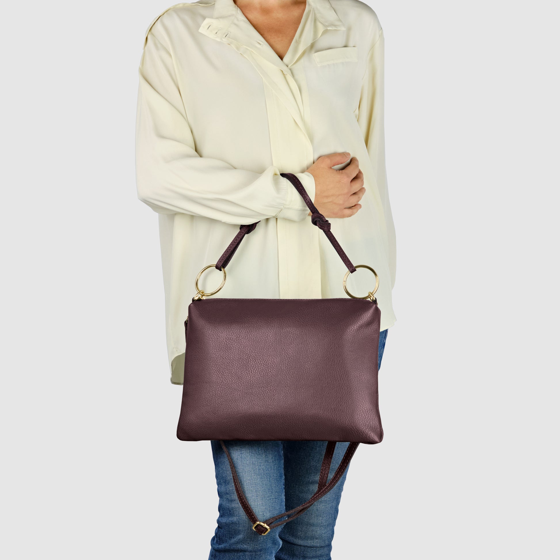 A person wearing a loose cream-colored blouse and blue jeans holds a structured Italian leather handbag with gold rings and a shoulder strap against a plain light gray background.