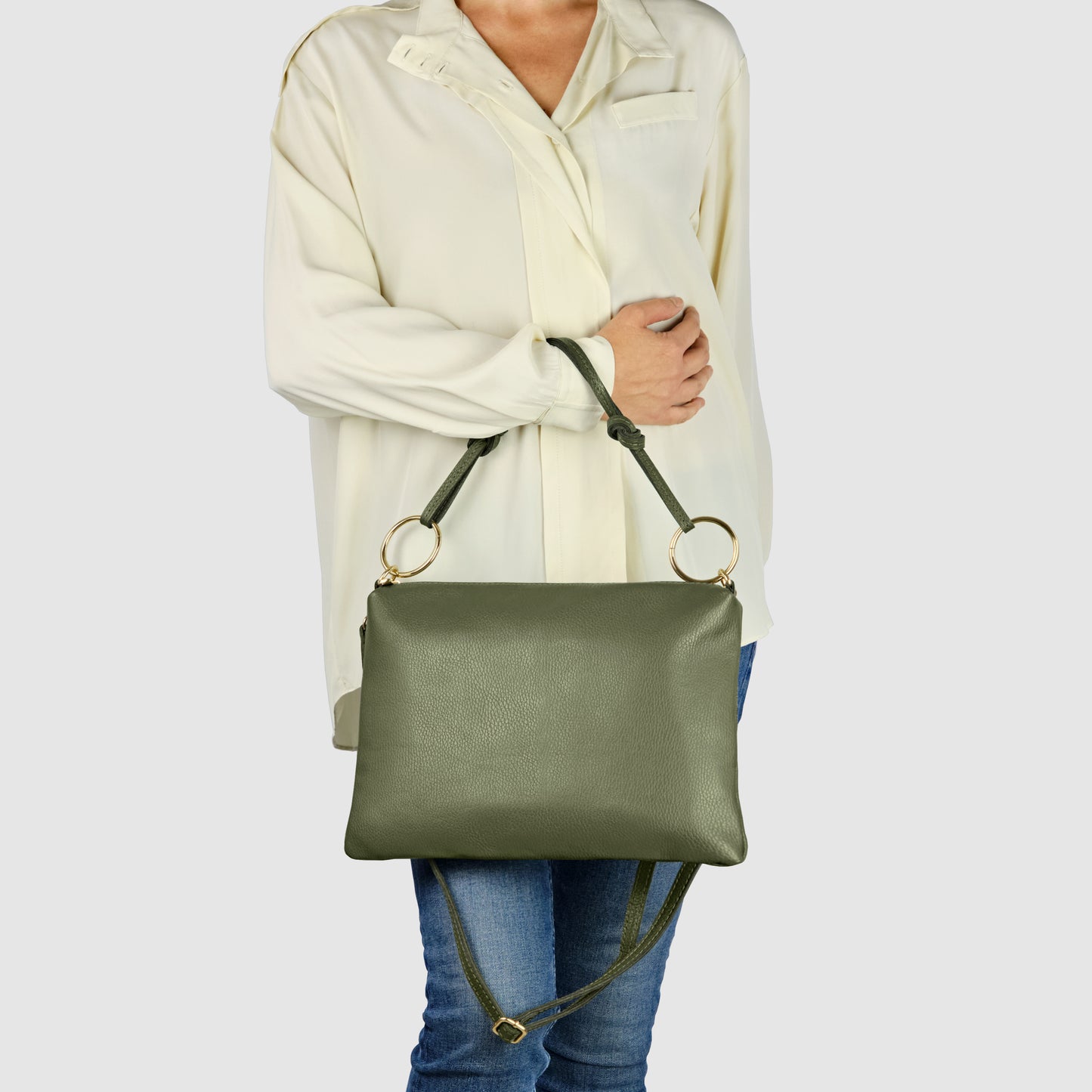 A person wearing a cream-colored blouse and blue jeans holds an Italian leather handbag with gold rings and an adjustable strap in front of their body.