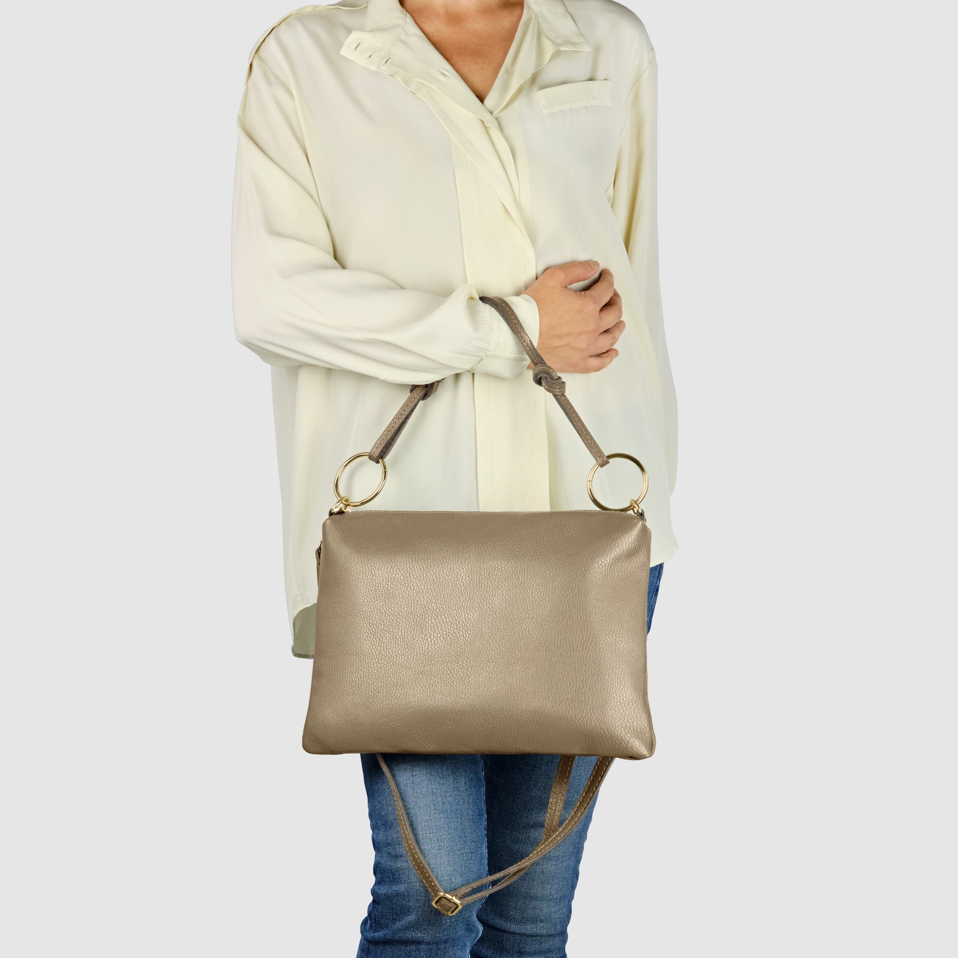 A person wearing a loose, cream-colored blouse and blue jeans holds an elegant crossbody bag—an Italian leather purse with gold rings and an adjustable strap—against a plain light background.