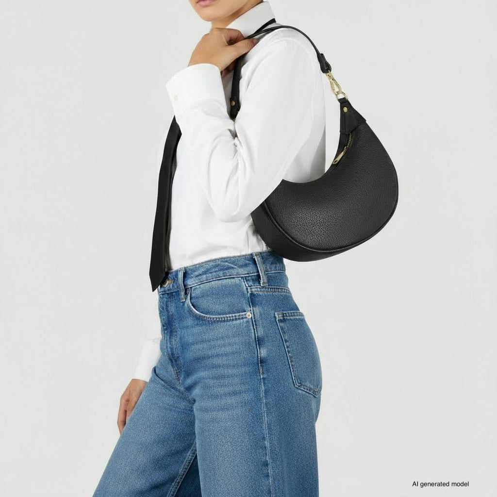 A person wearing a white shirt, black tie, and blue jeans holds a crescent-shaped handbag crafted from Italian calf leather against a plain light background. The model's face is partially out of frame.