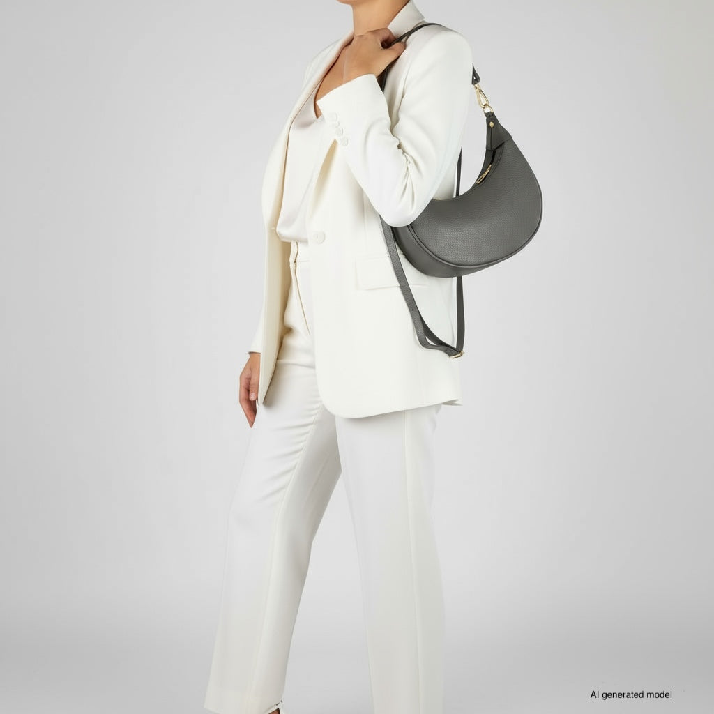 A person in a white suit holds a black crescent-shaped handbag crafted from Italian calf leather, standing against a plain gray background. The image features an AI-generated model.