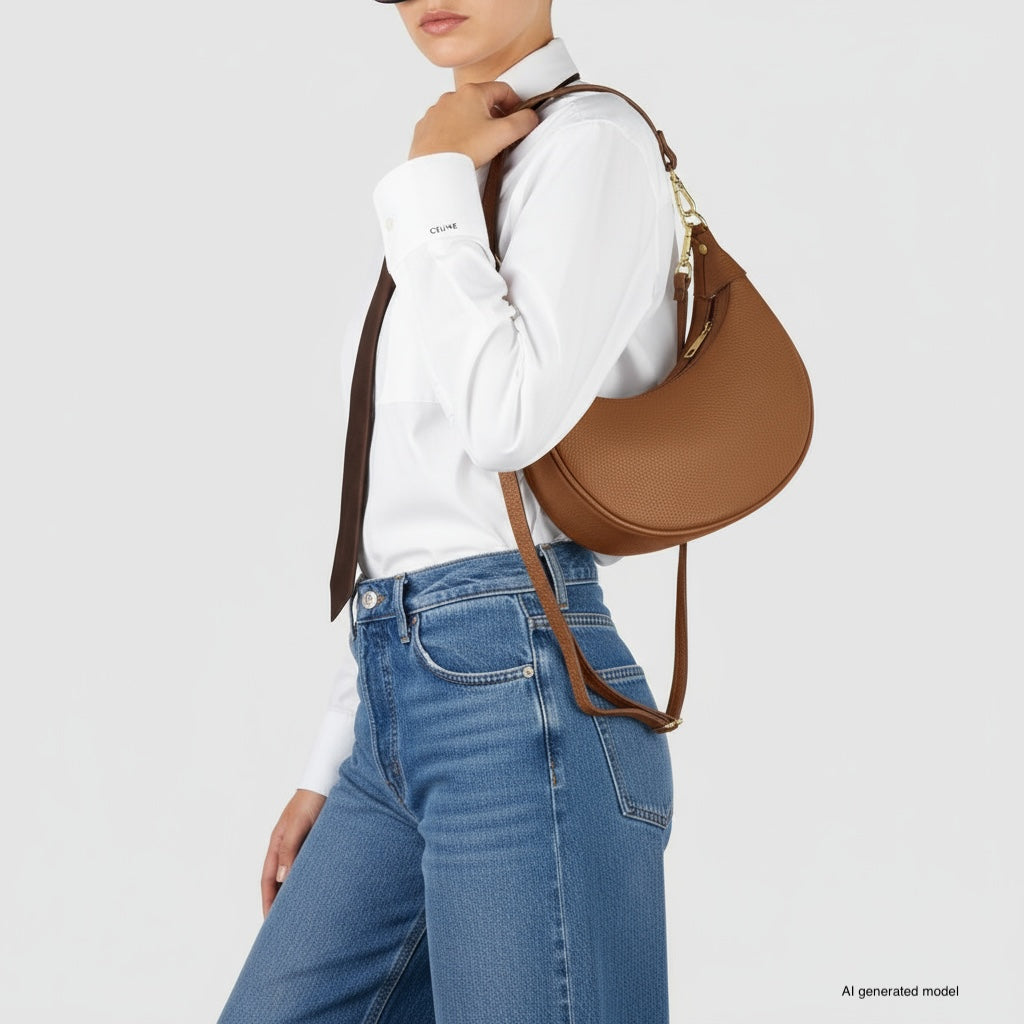 A person wearing a white shirt, dark tie, and blue jeans carries a crescent-shaped handbag crafted with Italian calf leather. The background is plain light gray.