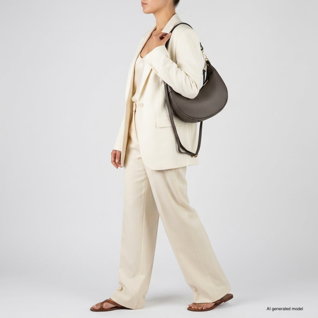 A woman in a cream-colored pantsuit and sandals carries a large, crescent-shaped handbag crafted with artisan craftsmanship. She stands sideways against a plain light background. AI generated model is noted in the image corner.
