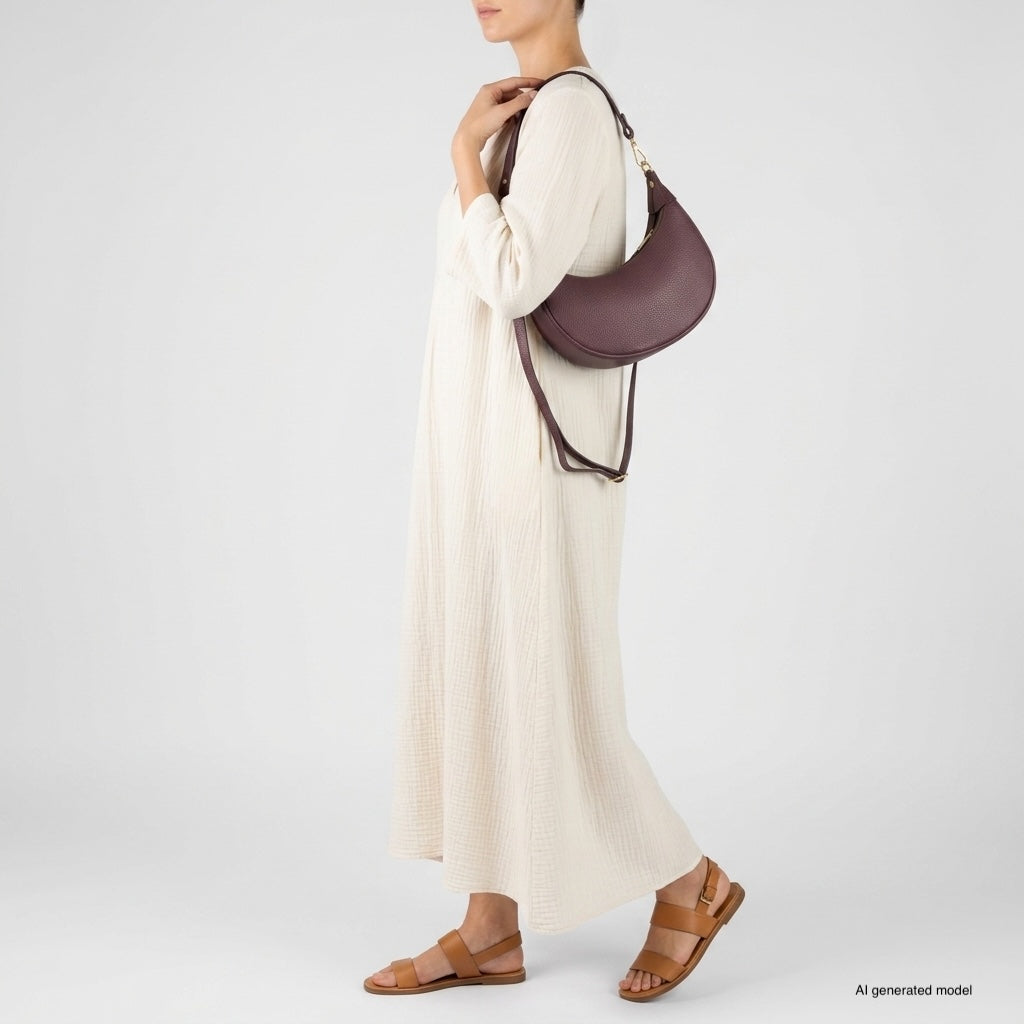 A person wearing a long, cream-colored dress and brown sandals stands sideways, holding a crescent-shaped handbag crafted from Italian calf leather. The background is plain and light-colored.