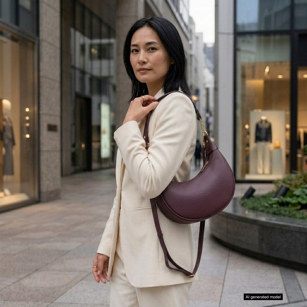 A woman in a cream suit stands in an urban outdoor shopping area, confidently holding a crescent-shaped handbag crafted with artisan craftsmanship. Storefronts and mannequins are visible in the background.