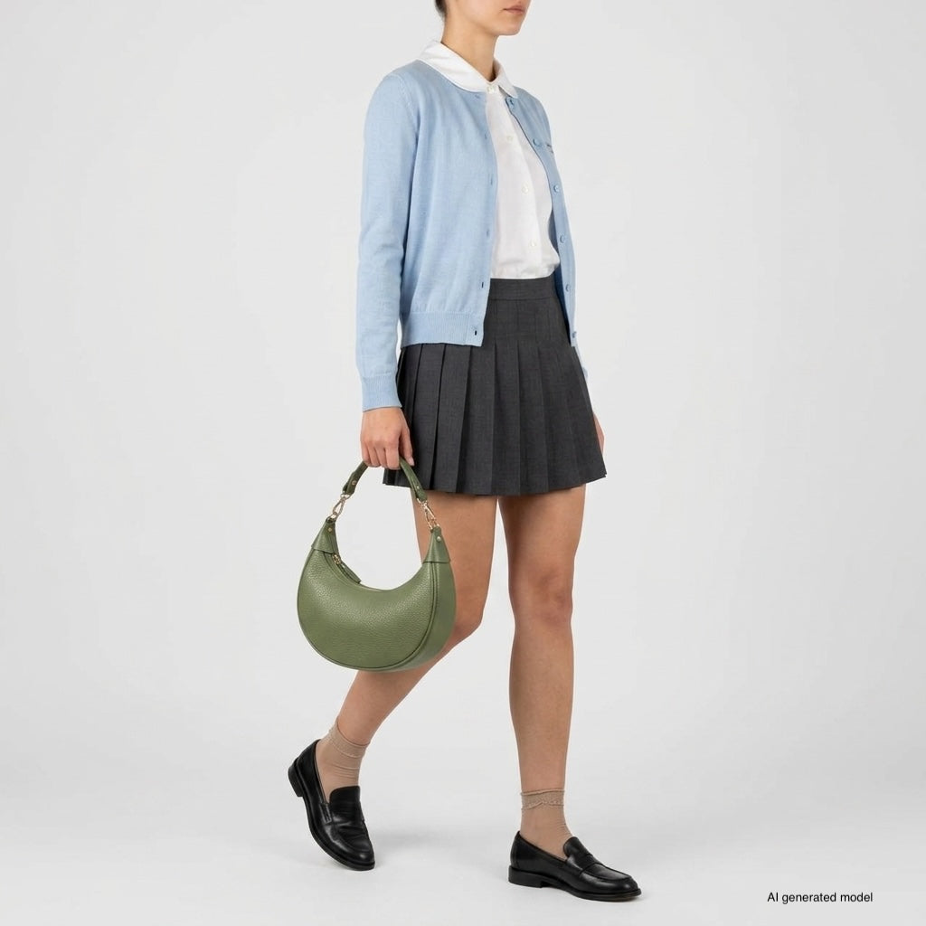 A person wearing a light blue cardigan, white shirt, gray pleated skirt, black loafers, and sheer socks holds a crescent-shaped handbag crafted with artisan craftsmanship. They are standing against a plain white background.