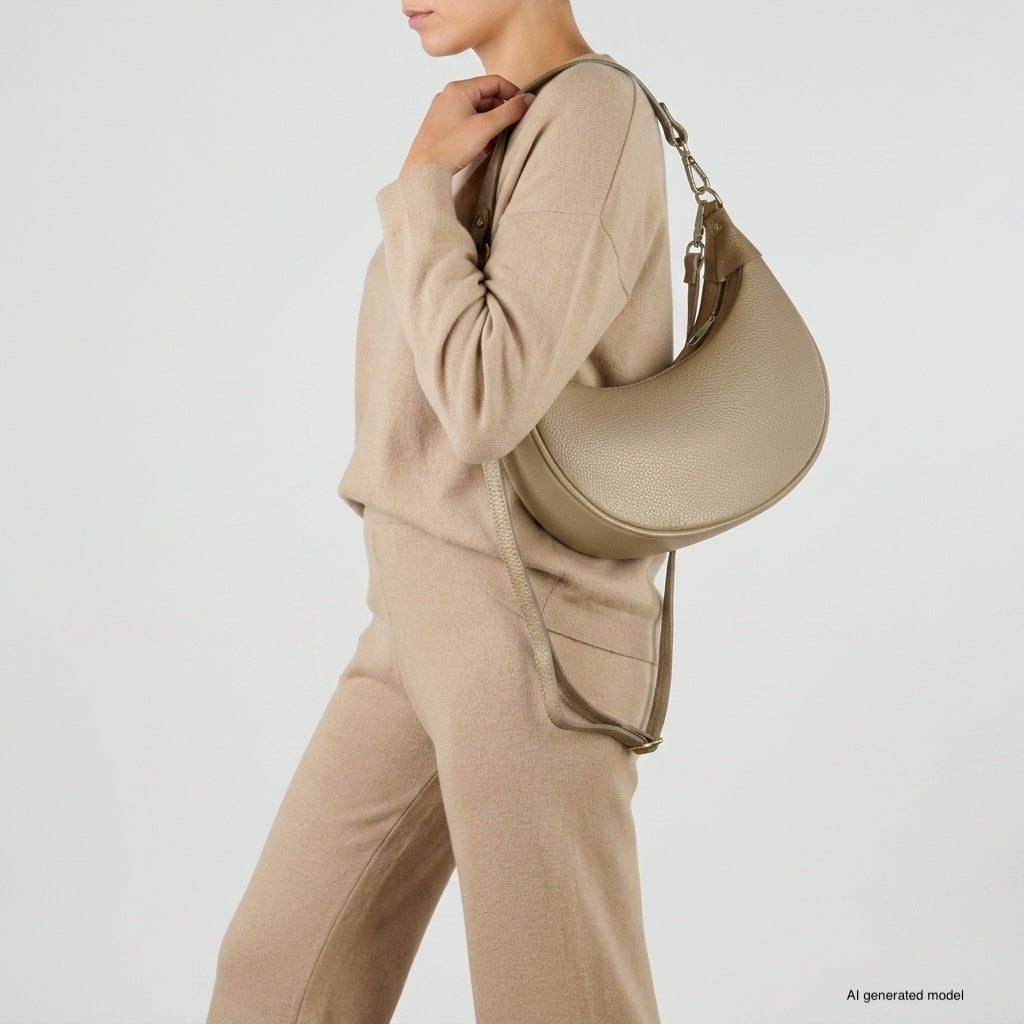 A woman wearing a beige knit sweater and pants holds a crescent-shaped handbag crafted from Italian calf leather, featuring exquisite artisan craftsmanship, posed against a plain light background.