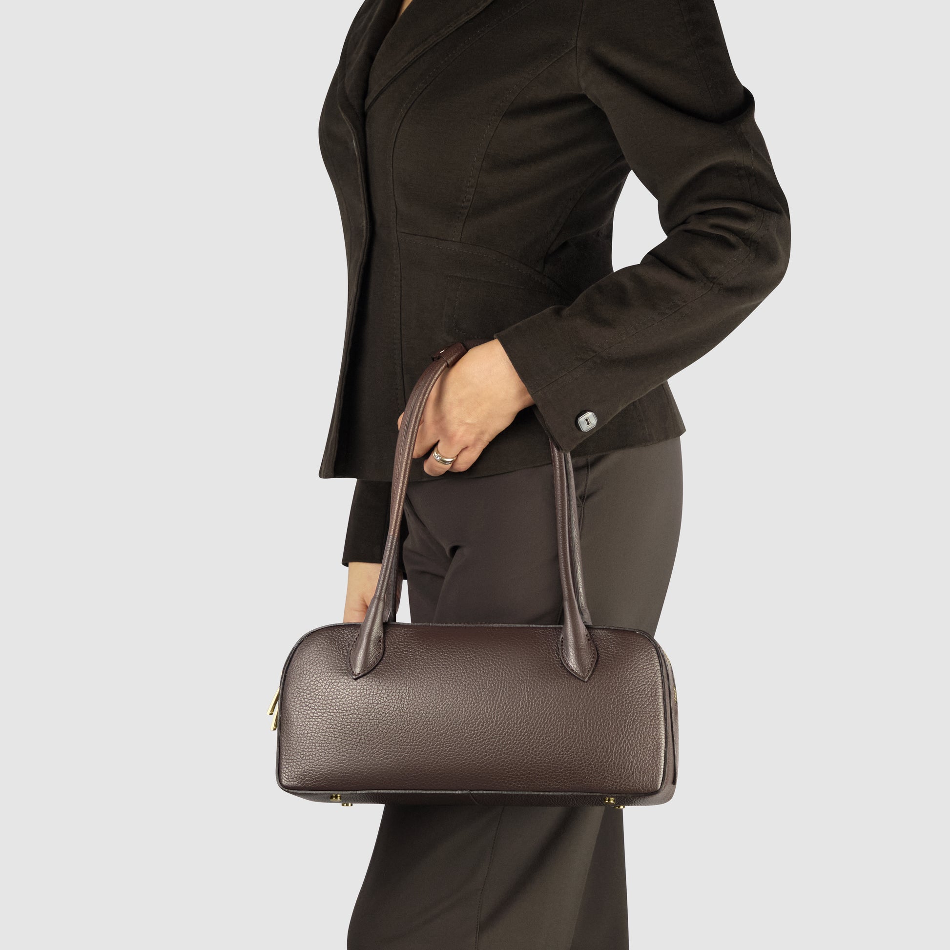 A woman in a dark blazer and pants holds an elegant Italian leather bag with two long handles, showcasing exquisite Florentine craftsmanship, against a light grey background.