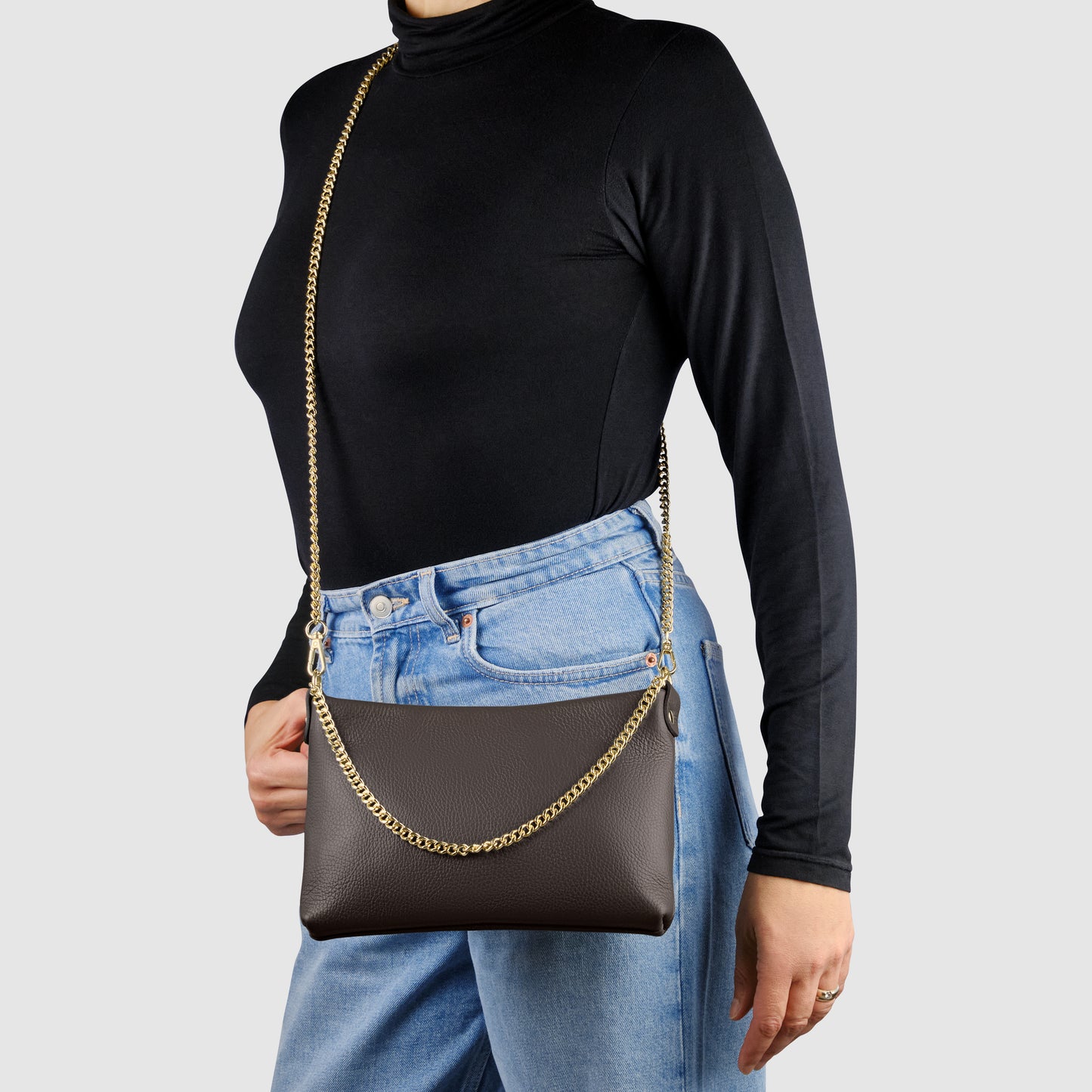 A person wearing a black turtleneck and blue jeans carries an Italian calf leather crossbody purse with gold chain straps. The background is plain light grey.