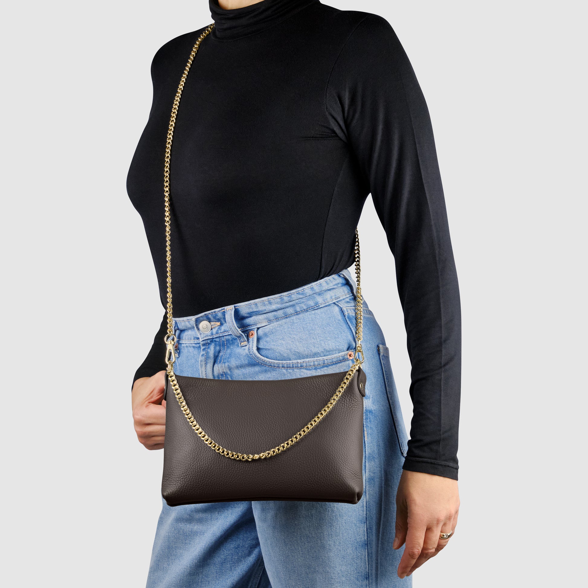 A person wearing a black turtleneck and blue jeans carries an Italian calf leather crossbody purse with gold chain straps. The background is plain light grey.