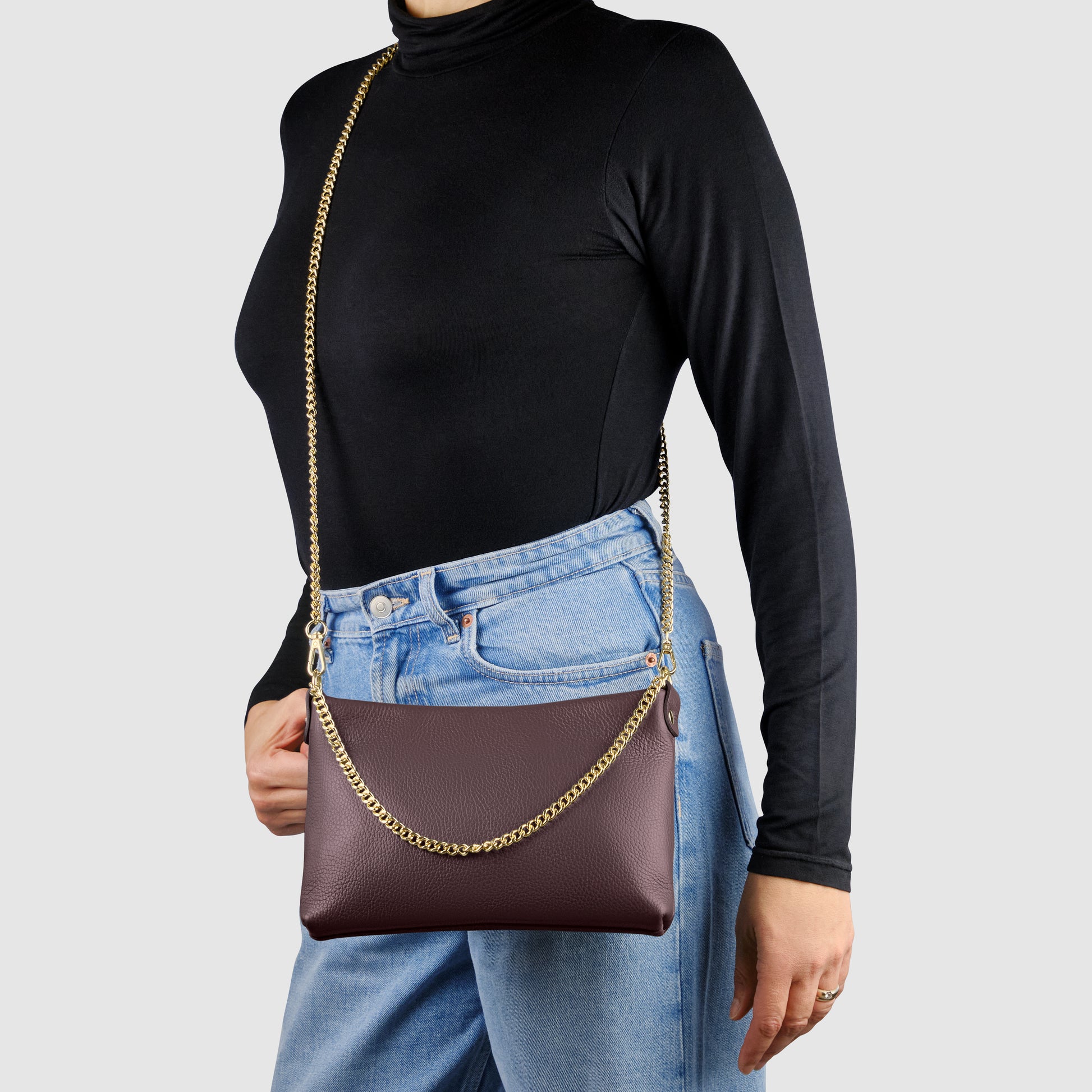 A person wearing a black turtleneck and blue jeans carries an Italian leather crossbody purse with a gold chain strap against a plain light gray background.