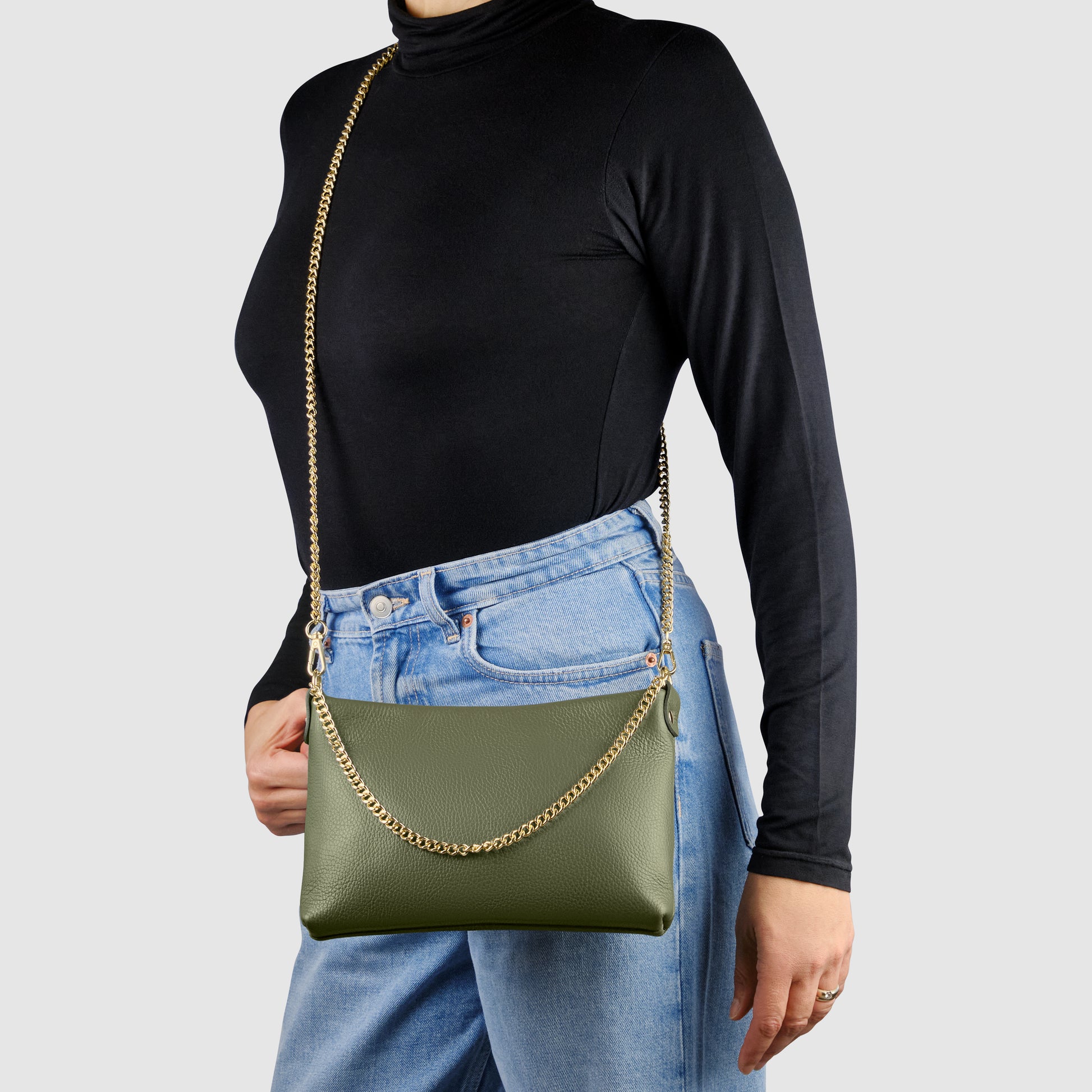A person wearing a black turtleneck and blue jeans holds an Italian leather crossbody purse with a gold chain strap, standing against a plain light gray background.