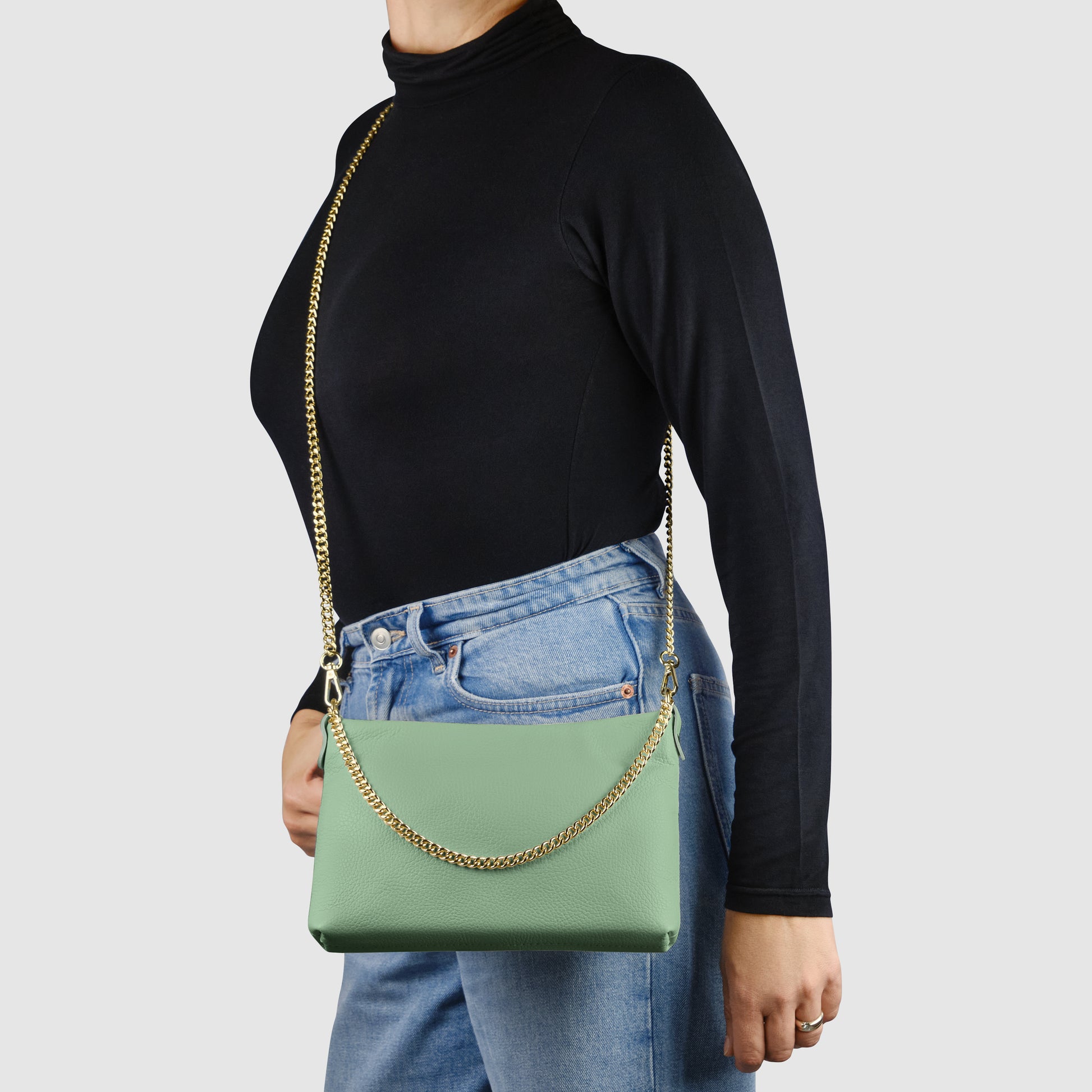 A person wearing a black long-sleeve top and blue jeans carries a handcrafted handbag with a gold chain strap over their shoulder. The light green Italian leather crossbody purse stands out against the plain, light-colored background.