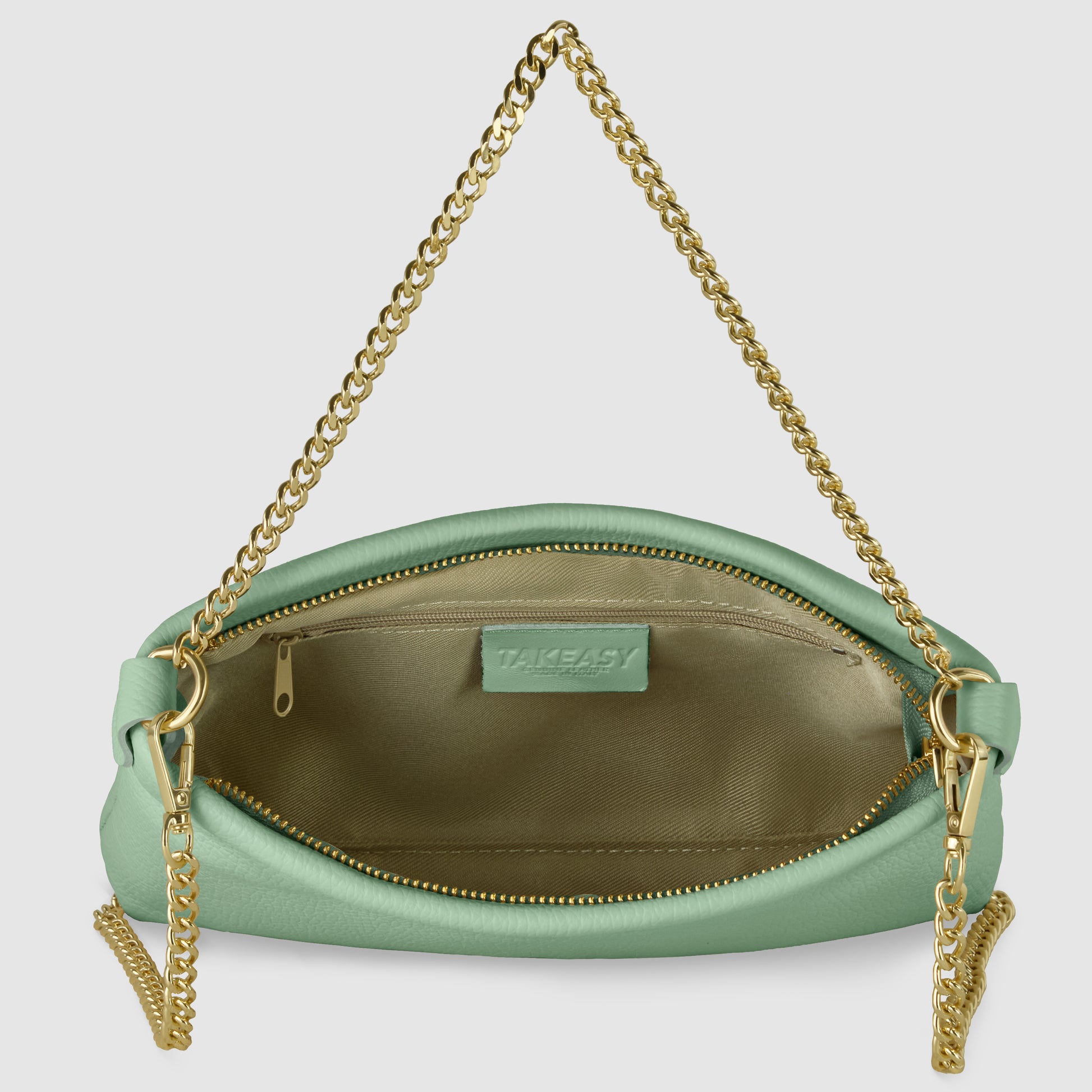 A small mint green handcrafted handbag with a gold chain strap is shown open, revealing a tan interior lining and a zippered pocket inside. The brand label TAKEASY is visible above the pocket.
