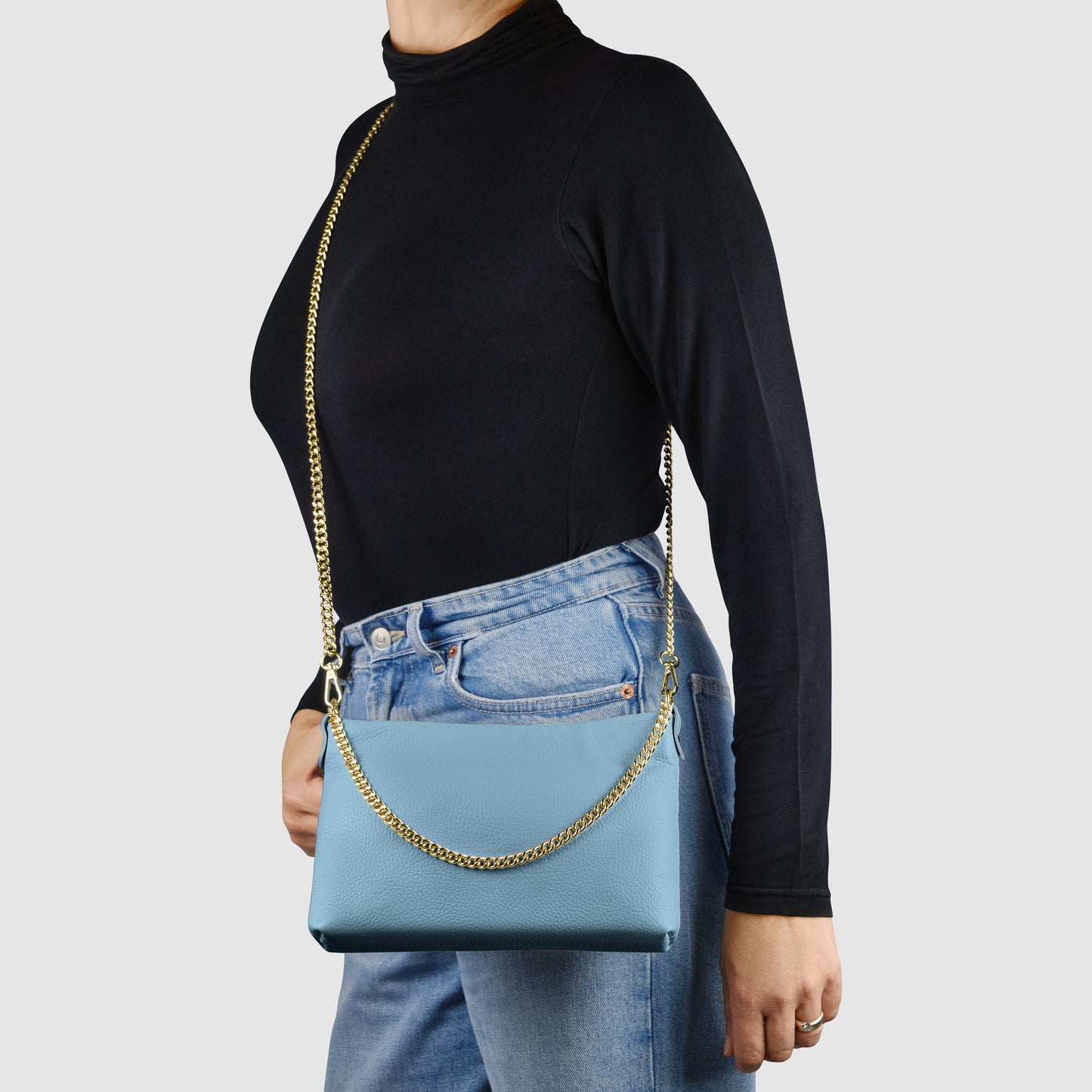 A person wearing a black long-sleeve top and blue jeans holds an Italian leather crossbody purse with a gold chain strap, against a plain light background.