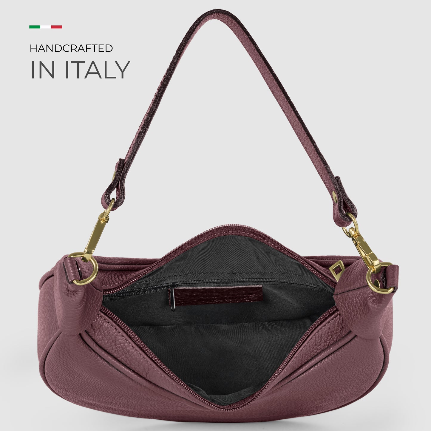 A crescent-shaped handbag in burgundy Italian calf leather with gold hardware and a short handle is shown open, displaying a black fabric-lined interior with a zip pocket. Text reads Handcrafted in Italy beside a small Italian flag.