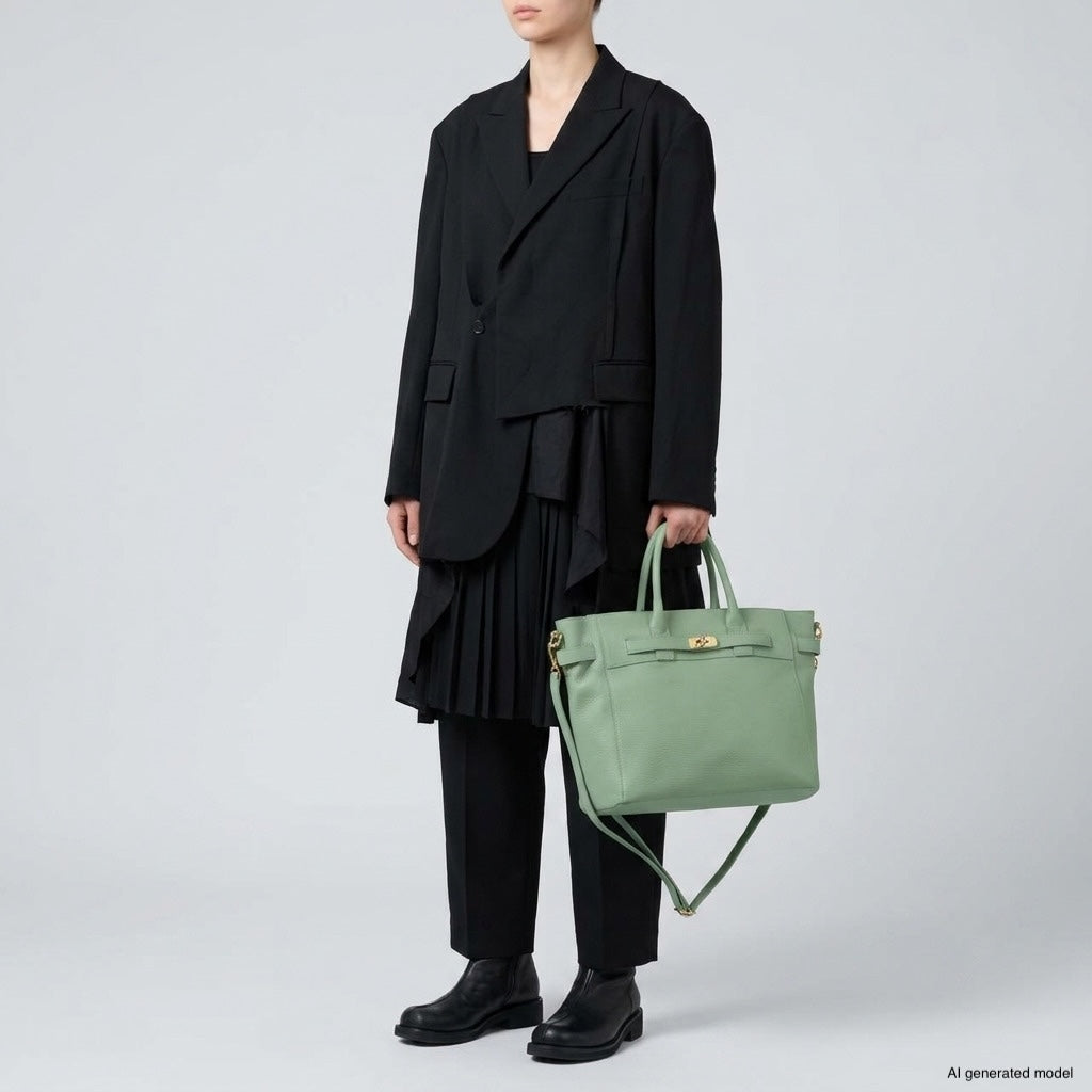 Person wearing a black oversized blazer, pleated skirt, black pants, and black shoes, holding a luxury tote bag with gold hardware, standing against a plain light gray background.
