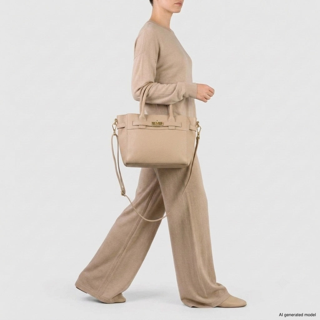 A person wearing a beige long-sleeve top and wide-leg pants walks while carrying a matching Italian leather handbag with a shoulder strap. The background is plain and light-colored.