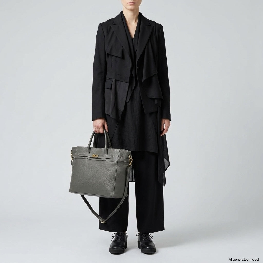 A person wearing a black layered jacket, wide-leg pants, and black shoes stands holding a luxury tote bag with a shoulder strap against a plain light background. The face is not visible.