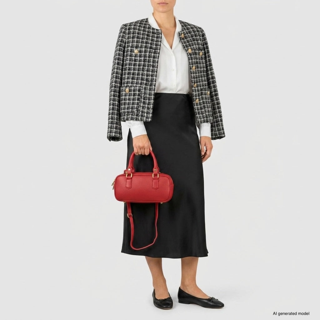 A woman in a white blouse, black midi skirt, and plaid jacket holds a minimalist structured bag. She wears black flats and stands against a plain light background.