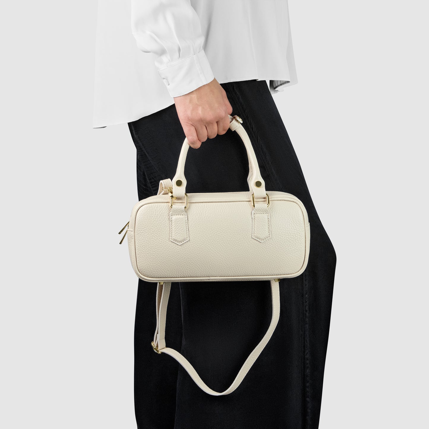 A person wearing a white shirt and black pants holds a small, rectangular, cream-colored minimalist handbag with both top handles and a shoulder strap.
