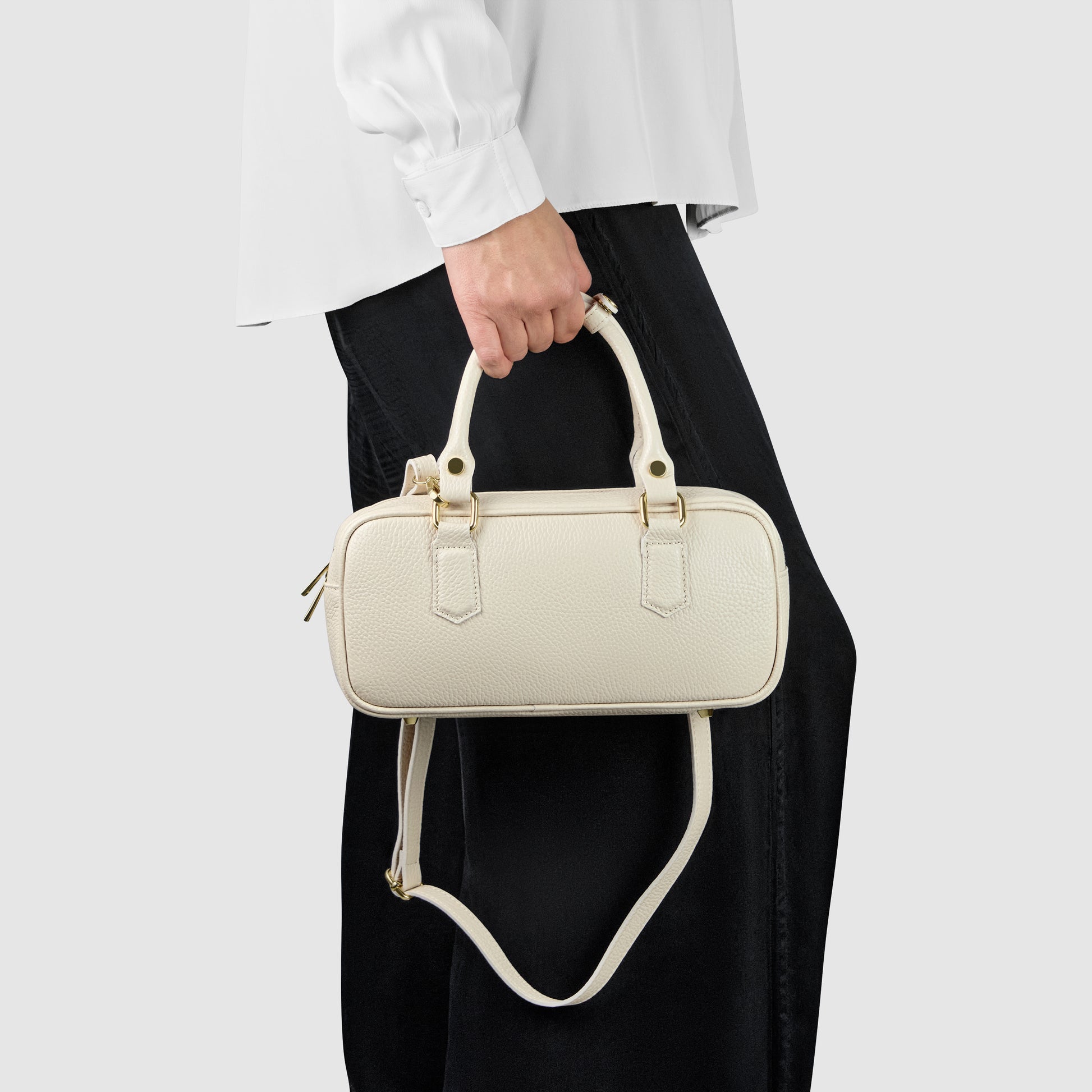 A person wearing a white shirt and black pants holds a small, rectangular, cream-colored minimalist handbag with both top handles and a shoulder strap.