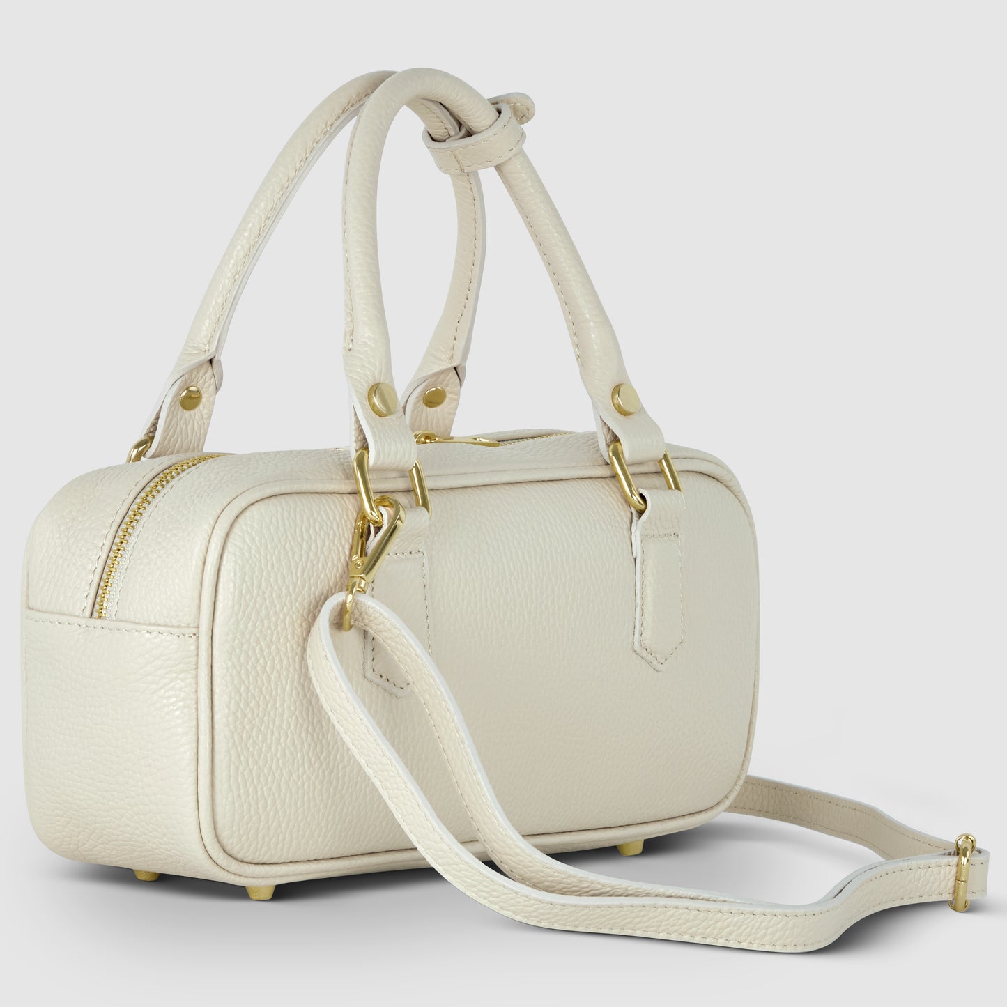 A small, rectangular, cream-colored Italian leather handbag with gold hardware, two short handles, and a detachable shoulder strap. The bag sits on four metal feet and features a pebbled finish with a top zipper closure.