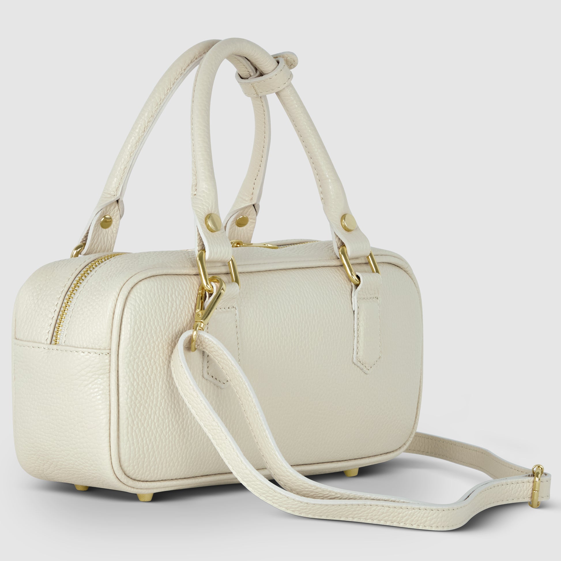 A small, rectangular, cream-colored Italian leather handbag with gold hardware, two short handles, and a detachable shoulder strap. The bag sits on four metal feet and features a pebbled finish with a top zipper closure.