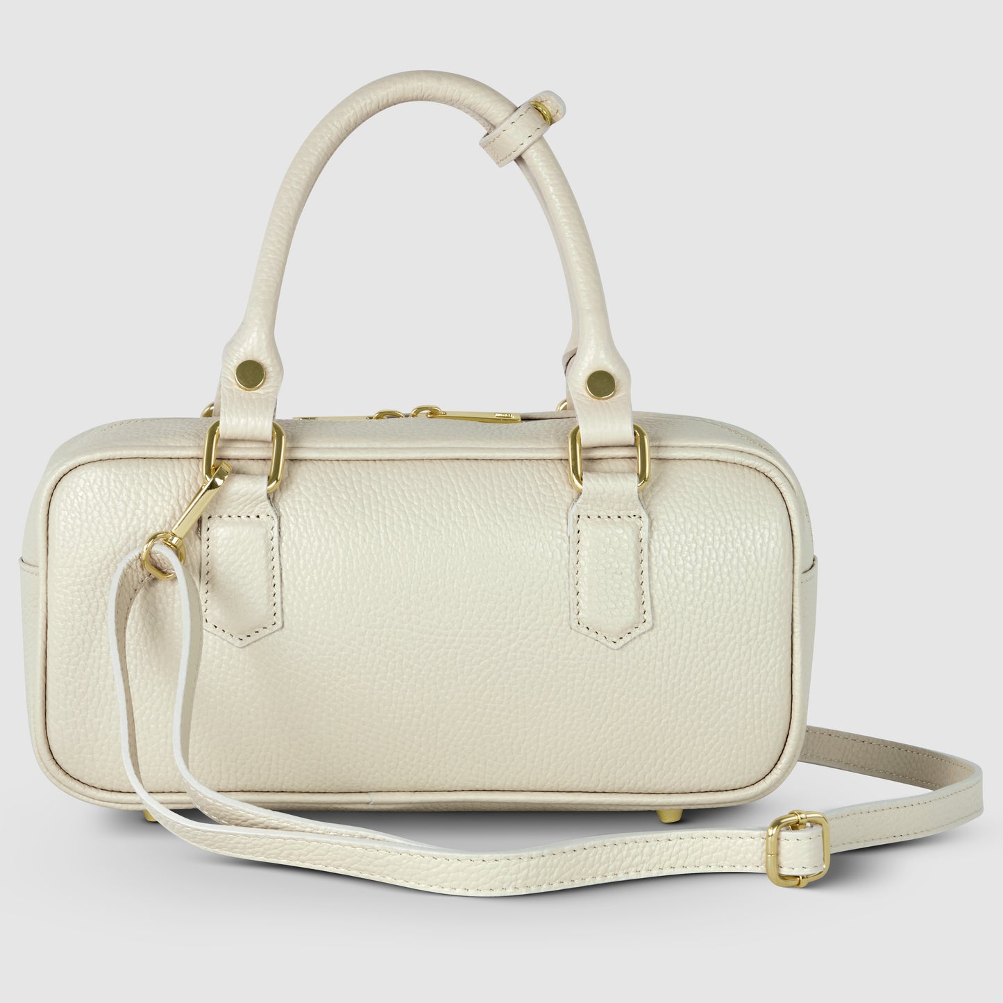 A small, rectangular, cream-colored minimalist handbag crafted from pebbled leather, featuring gold hardware, two short handles, and a detachable adjustable shoulder strap, displayed against a plain light gray background.