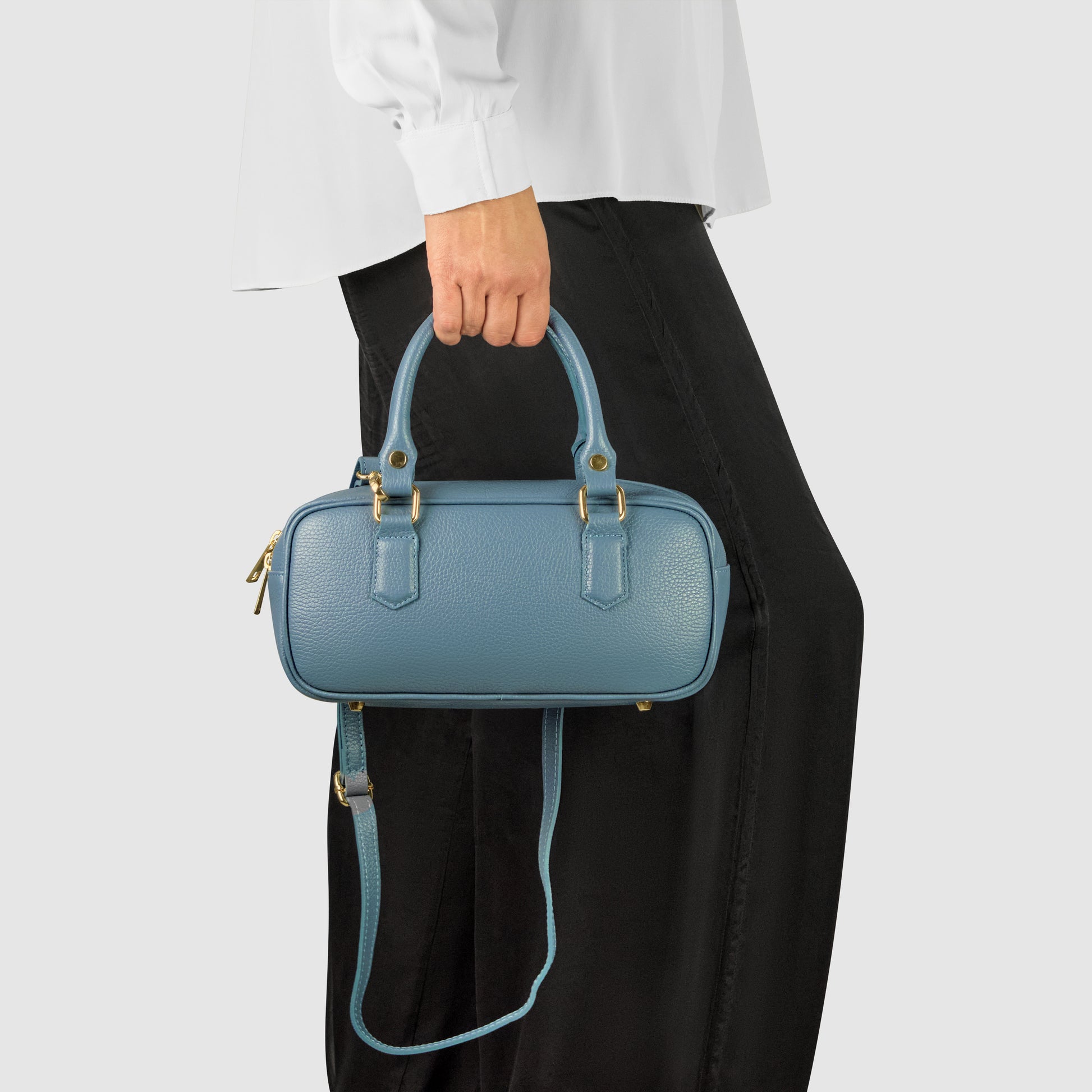 A person in a white long-sleeve shirt and black skirt holds a small, rectangular blue minimalist handbag with gold hardware and a detachable shoulder strap against a plain background.