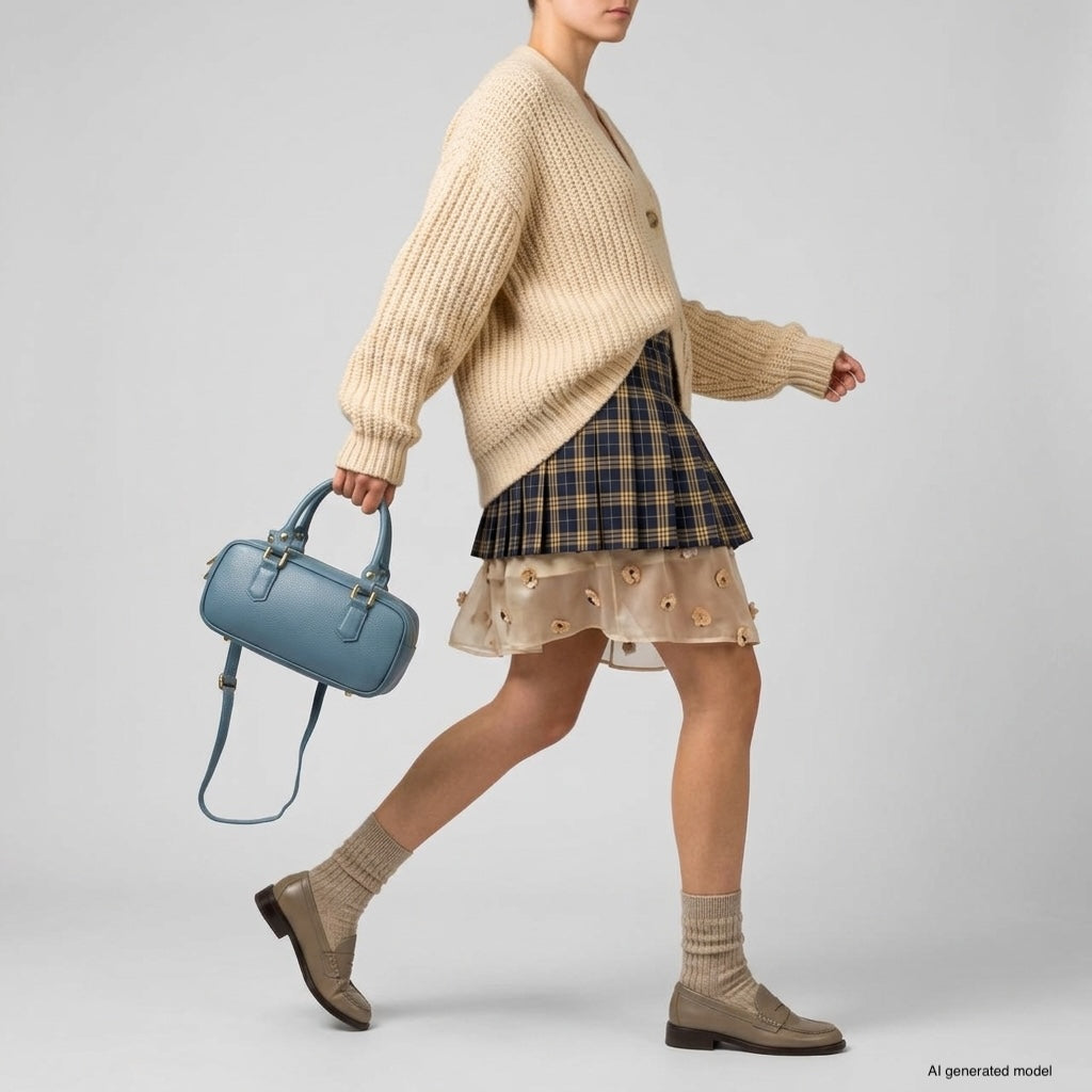 A person wearing a beige chunky knit cardigan, plaid skirt with a sheer overlay, beige socks, and tan shoes walks while holding a minimalist structured bag in light blue against a plain background.