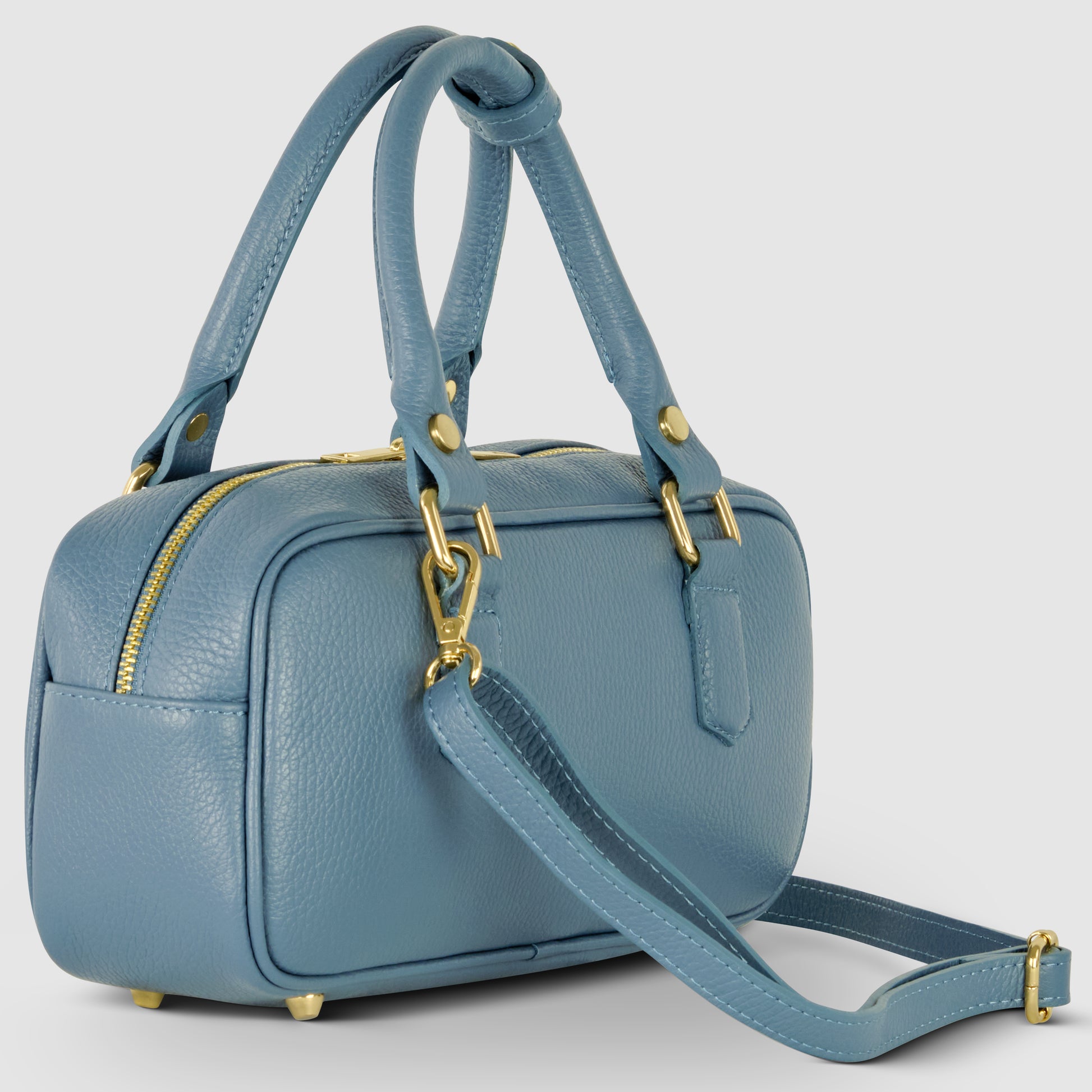 A small, rectangular blue Italian leather handbag with gold hardware, double handles, a detachable shoulder strap, and a zippered top. The pebbled leather bag sits on four small metal feet against a light background.