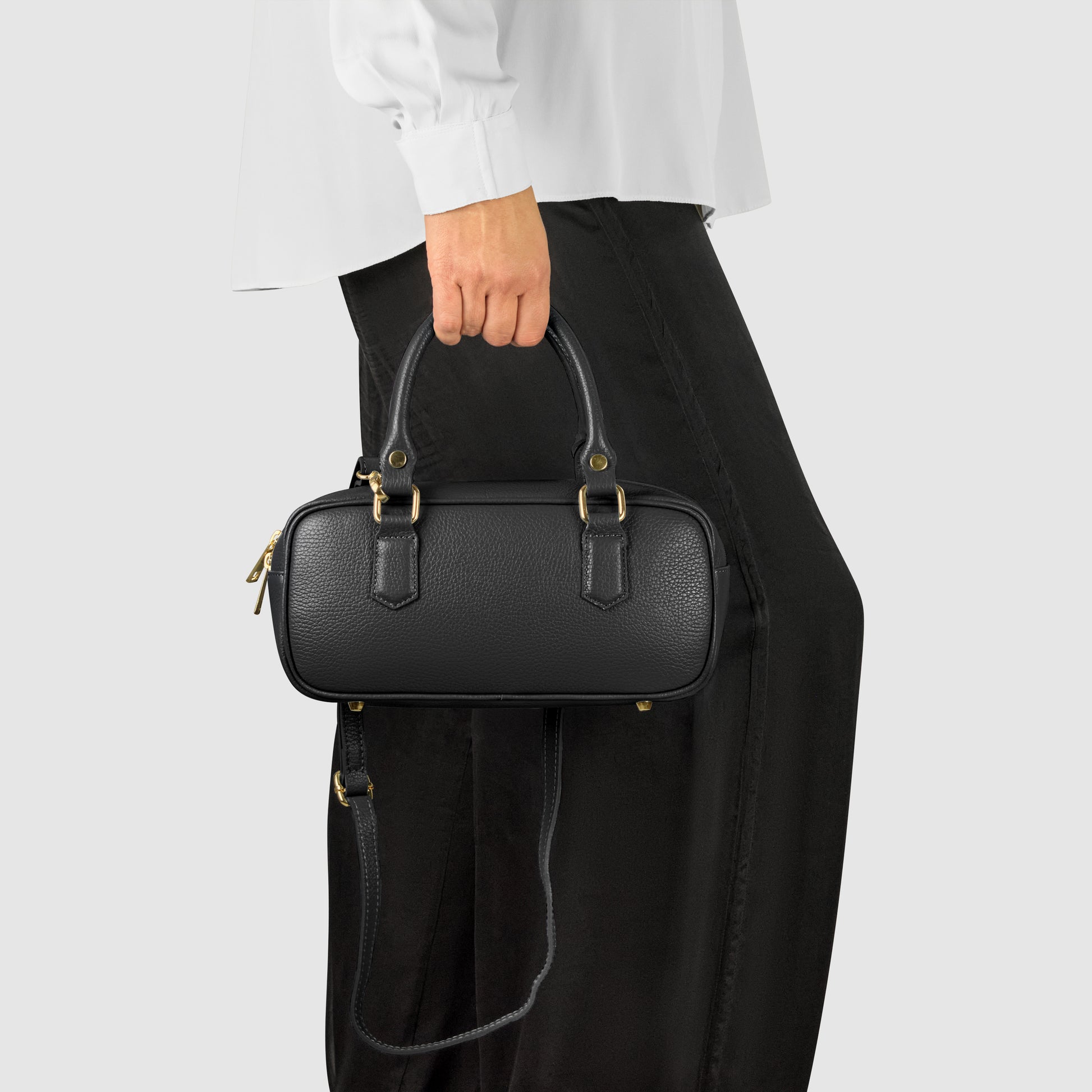 A person wearing a white long-sleeve shirt and black skirt is holding a small minimalist handbag with gold hardware and a shoulder strap against a plain light gray background.