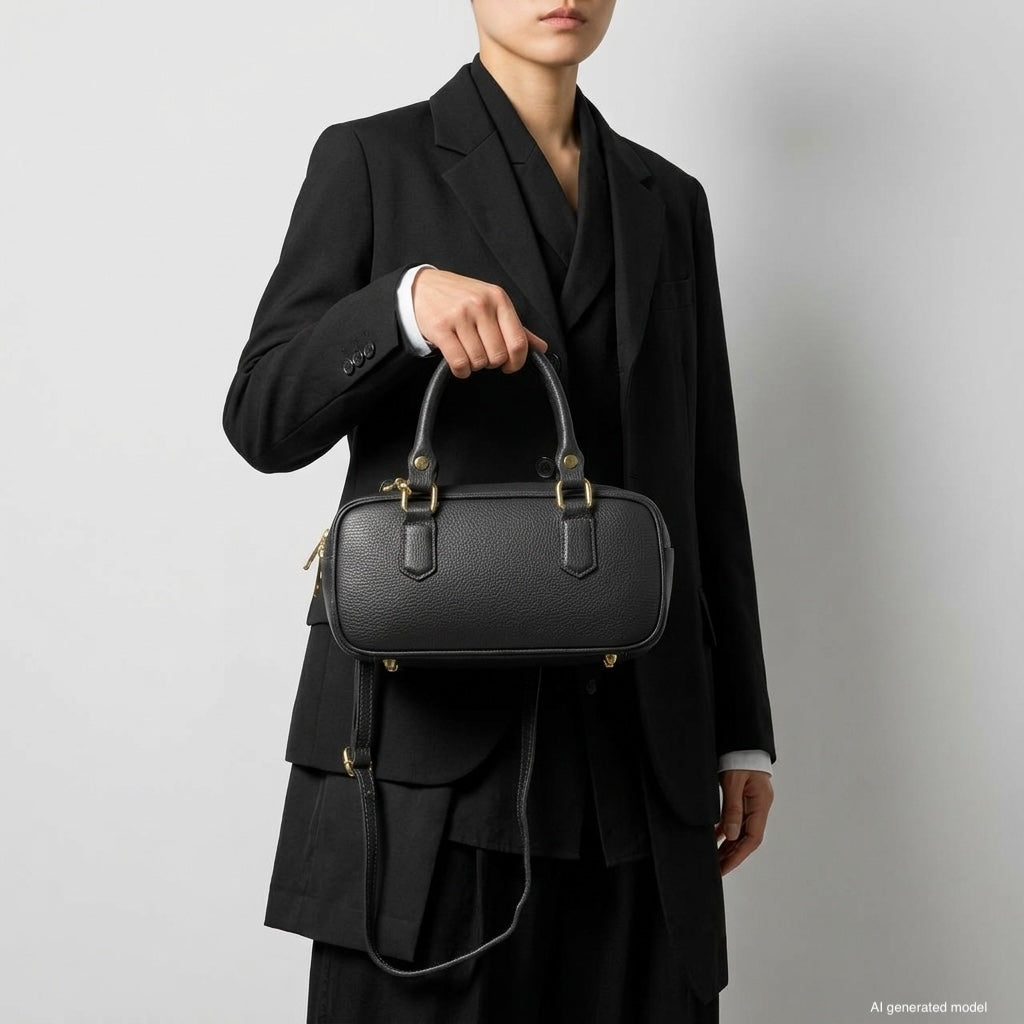 A person wearing a black suit holds a minimalist structured bag with gold hardware and a detachable strap against a plain light background. The person's face is partially out of frame.