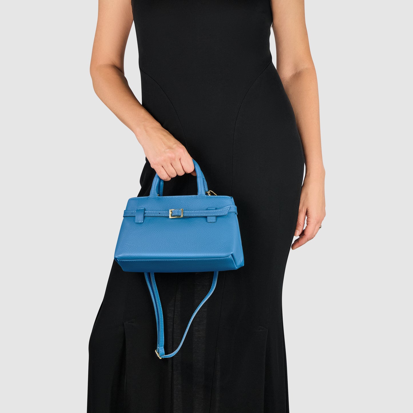 A woman wearing a sleeveless black dress holds a small blue Italian leather handbag with a top handle and shoulder strap, embodying minimalism against a plain light background.