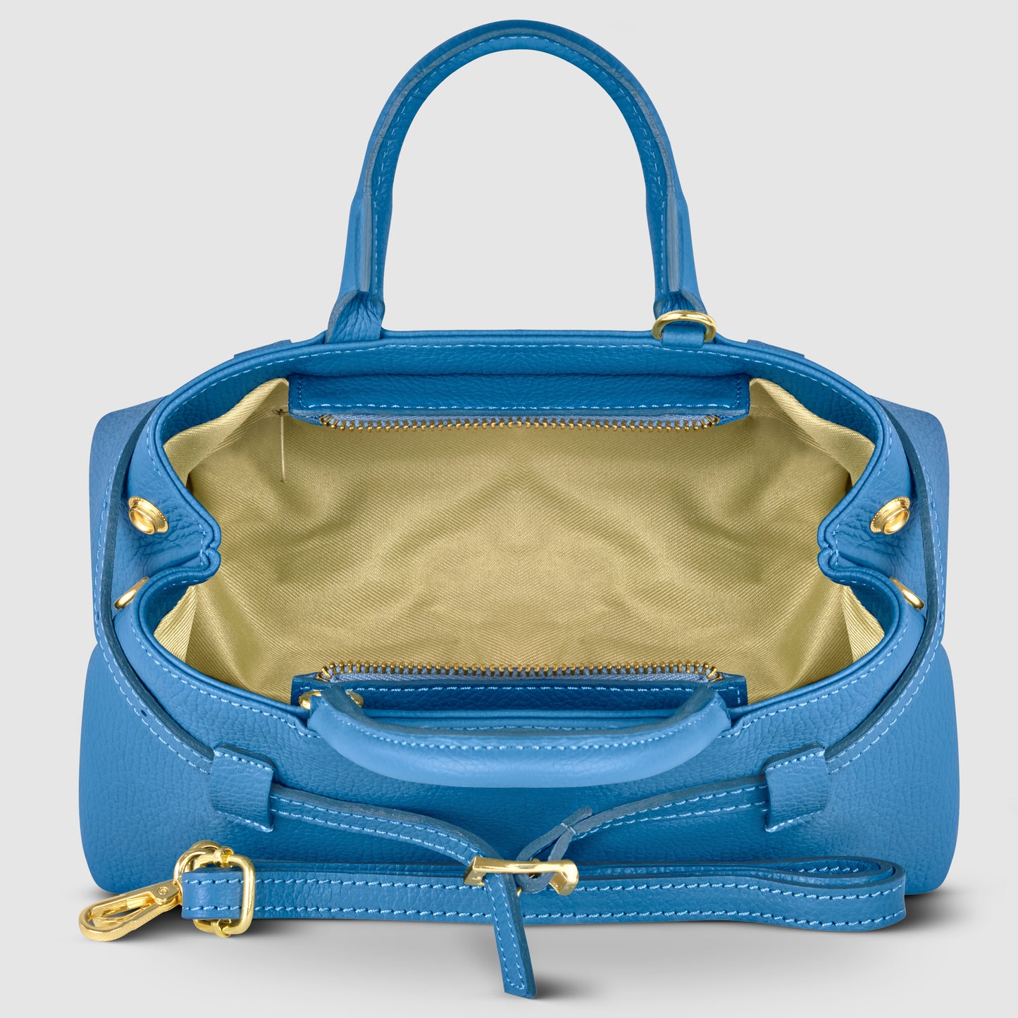 A blue Italian leather handbag with gold hardware is shown open, revealing a spacious beige fabric-lined interior with a zippered pocket and an adjustable shoulder strap attached; this minimalism bag is handmade in Italy.