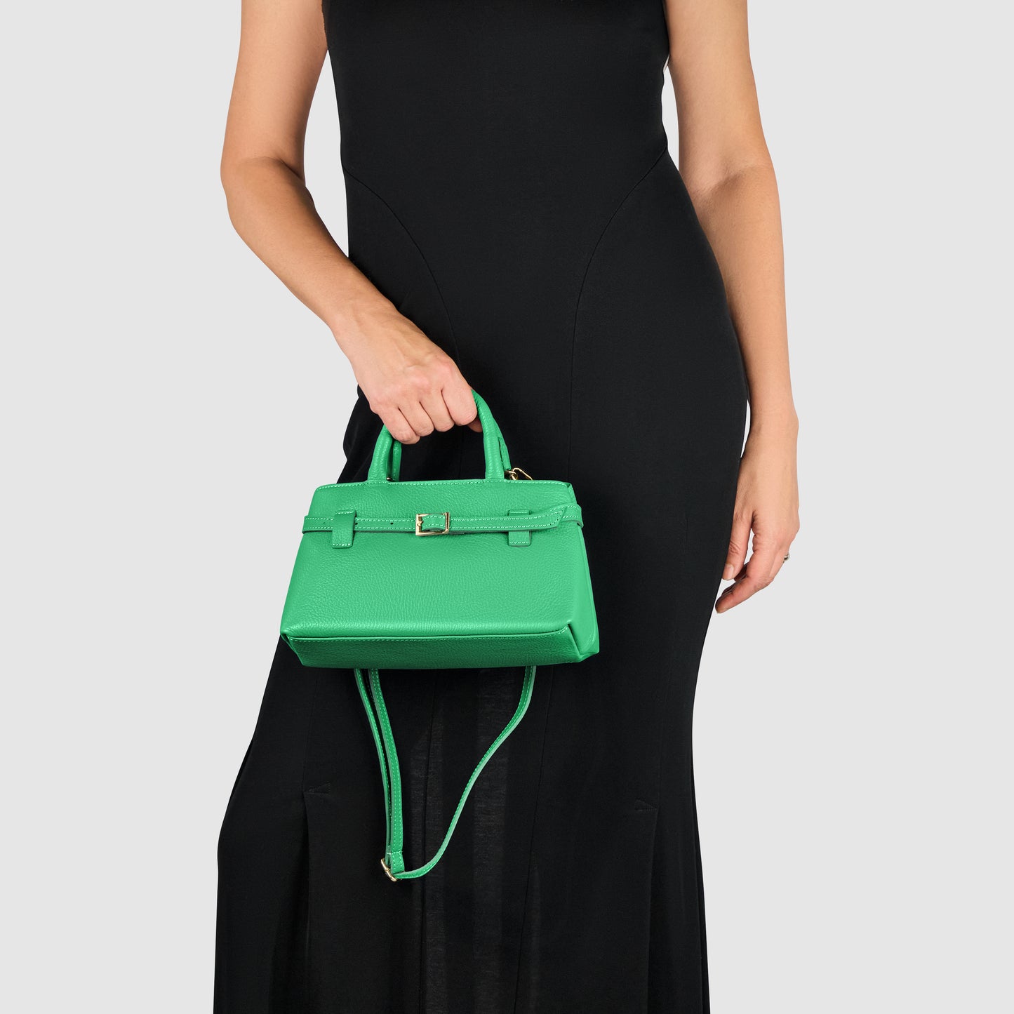 A woman in a sleeveless black dress holds a minimalist handbag—an elegant small green accessory with a top handle and shoulder strap—against a plain light gray background.