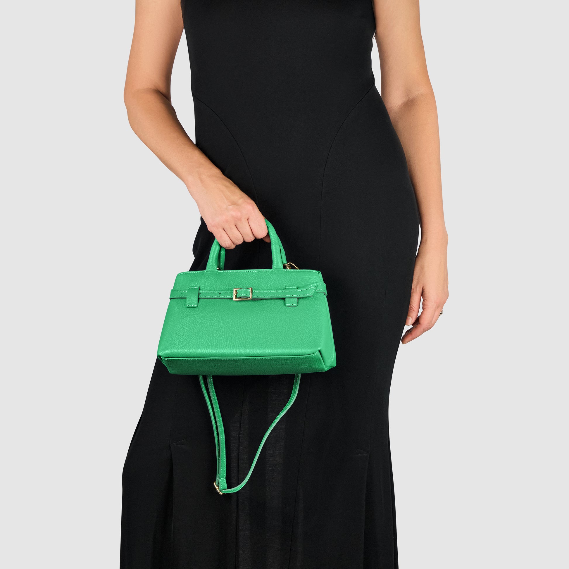 A woman in a sleeveless black dress holds a minimalist handbag—an elegant small green accessory with a top handle and shoulder strap—against a plain light gray background.