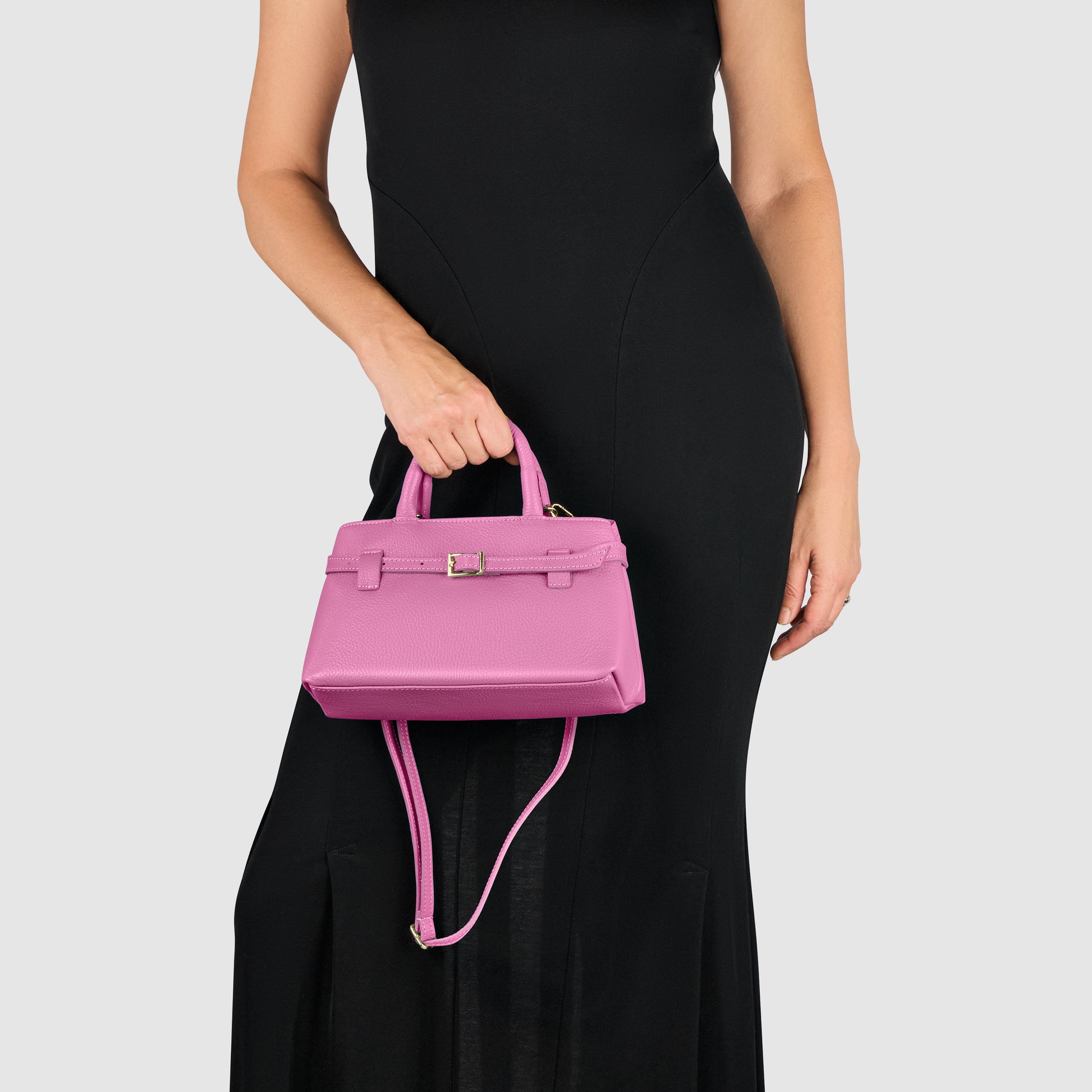 A woman in a sleeveless black dress holds a small pink minimalist handbag with a top handle and shoulder strap against a plain light background.
