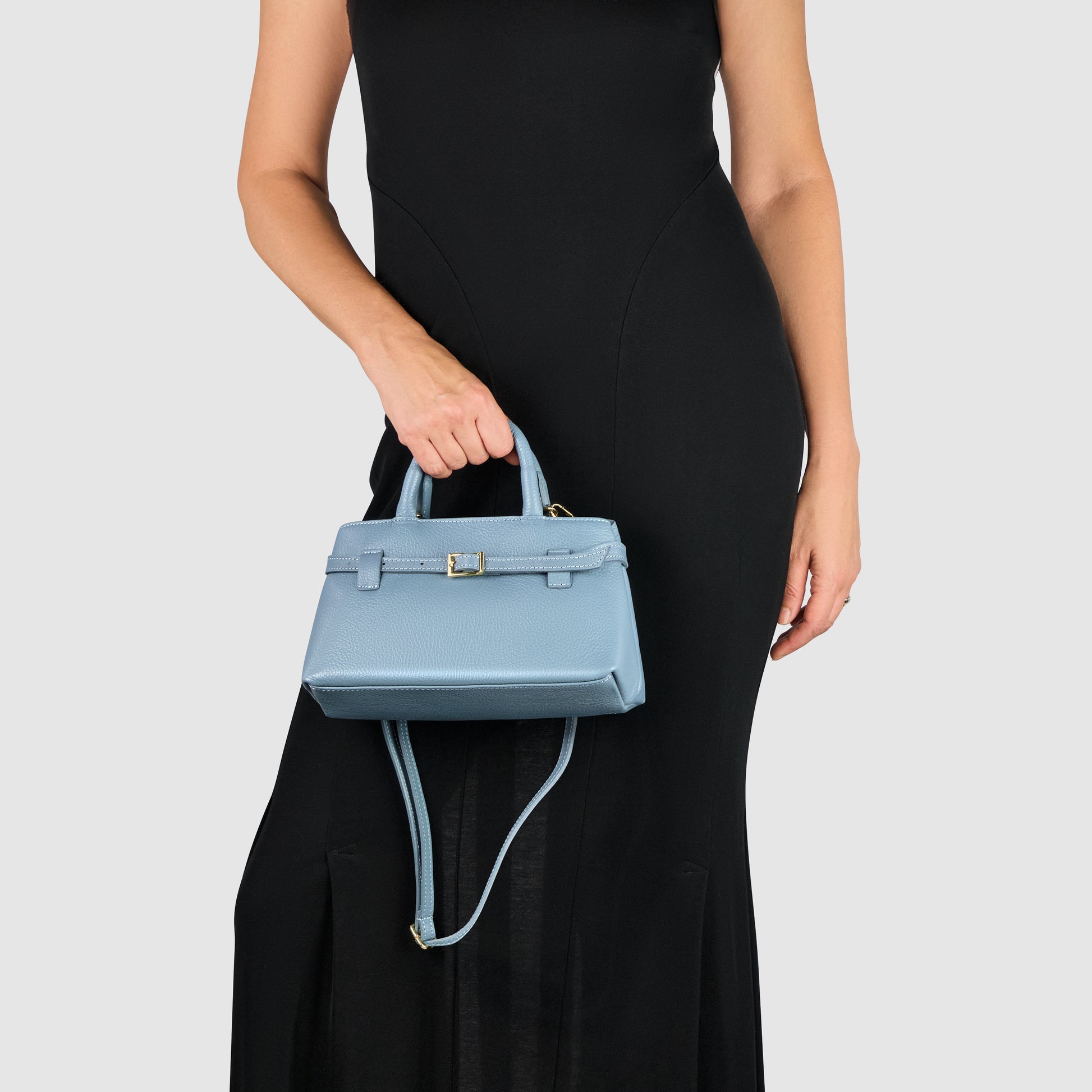 A woman in a sleeveless black dress holds a small, structured minimalist handbag with a shoulder strap in her left hand against a plain light background.