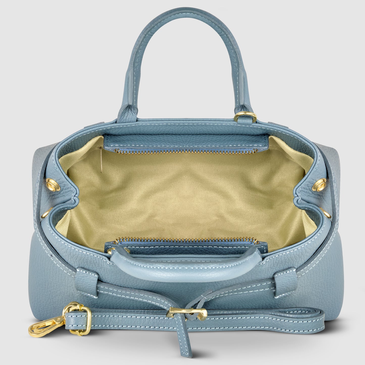 A light blue Italian leather handbag with gold hardware, open to reveal a spacious, light beige interior and a zippered pocket. This minimalist handbag features two handles and an adjustable shoulder strap, handmade in Italy.