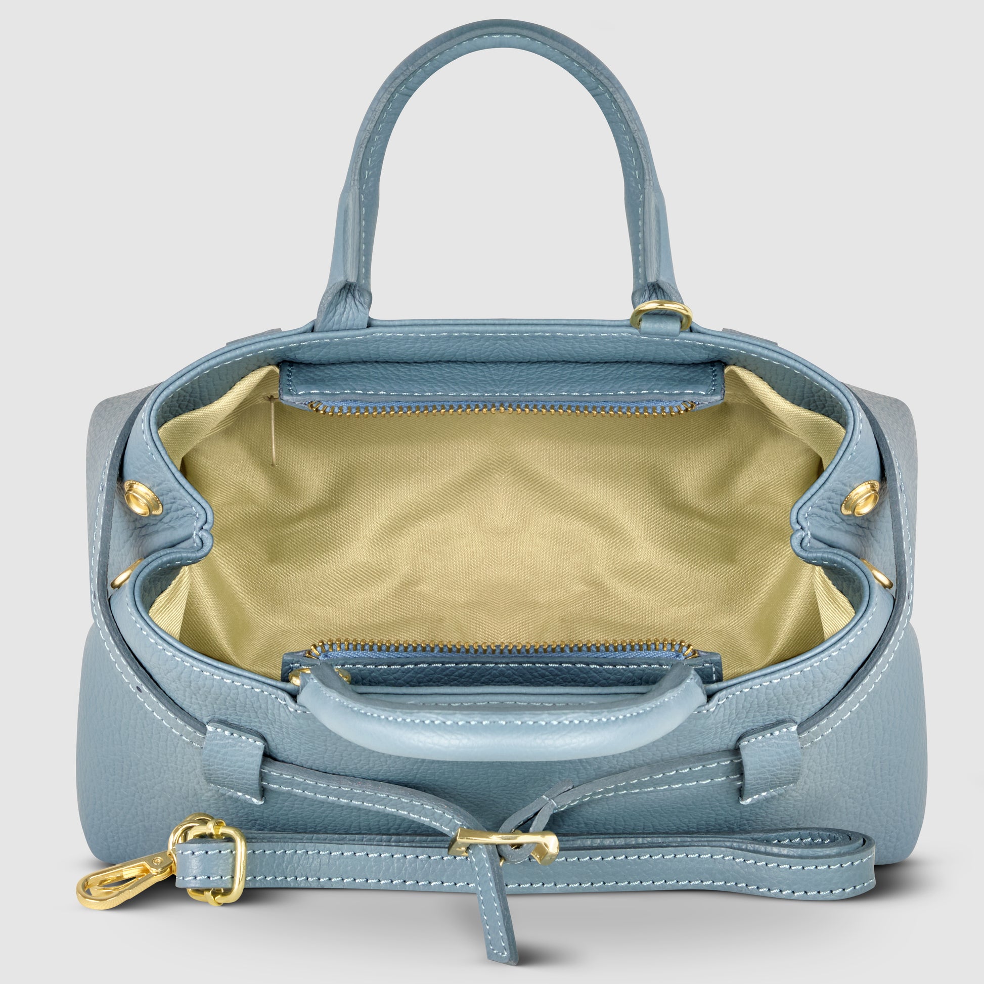 A light blue Italian leather handbag with gold hardware, open to reveal a spacious, light beige interior and a zippered pocket. This minimalist handbag features two handles and an adjustable shoulder strap, handmade in Italy.