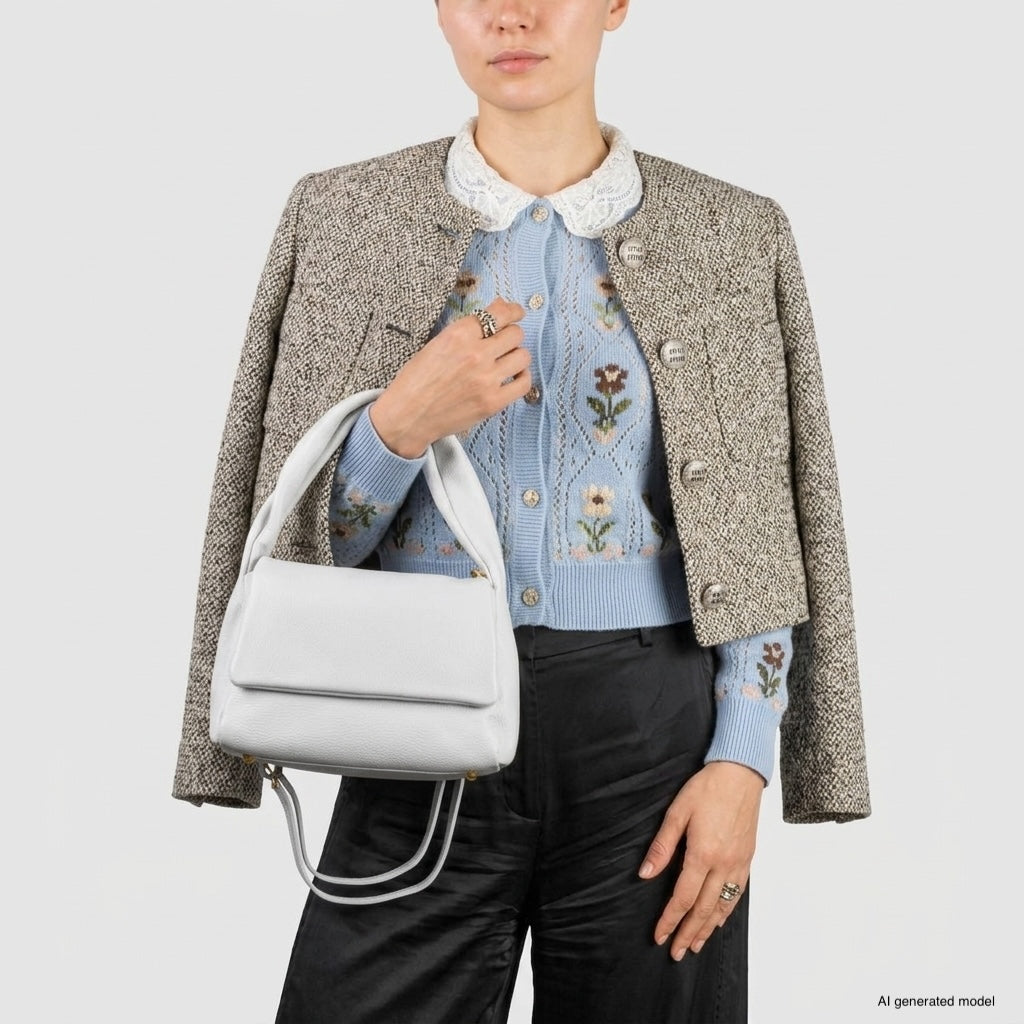 A person wearing a tweed jacket over a blue floral sweater and black pants holds a medium rectangular handbag made in Italy. The background is plain and light-colored.