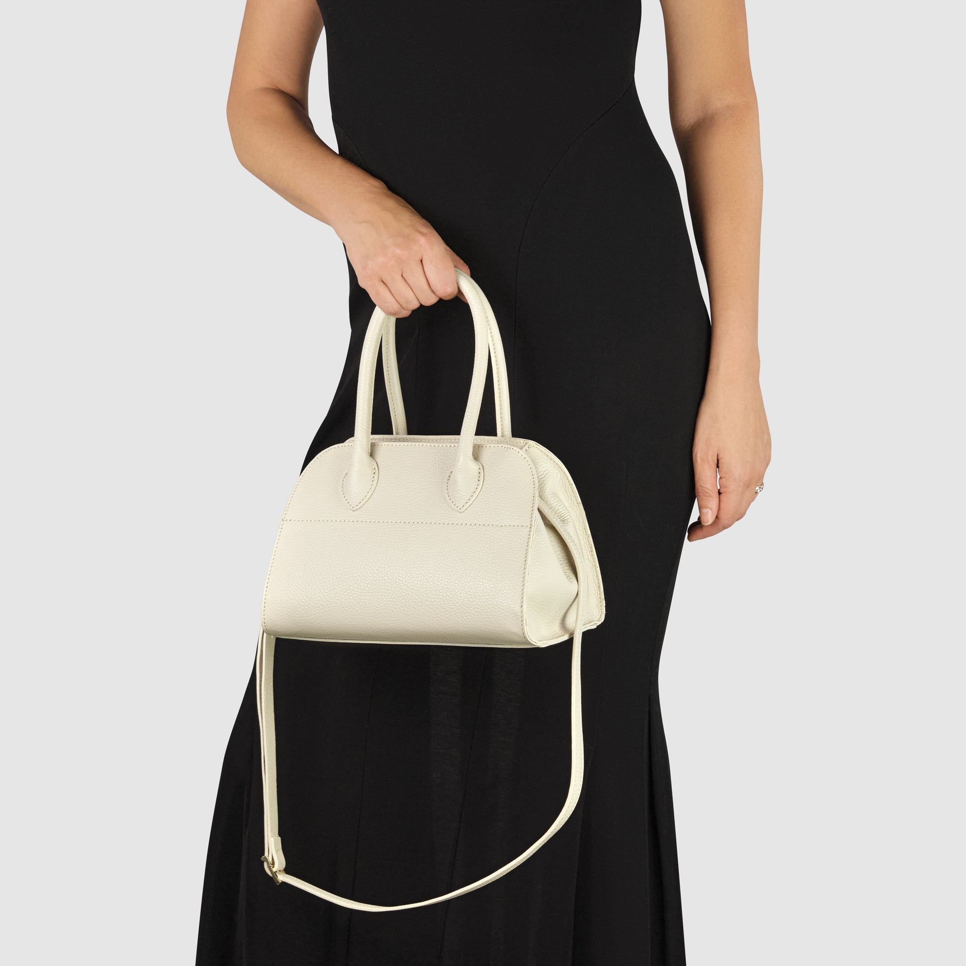 A woman in a black sleeveless dress holds a cream-colored Italian leather handbag with both an upper handle and a shoulder strap, standing against a plain light background.