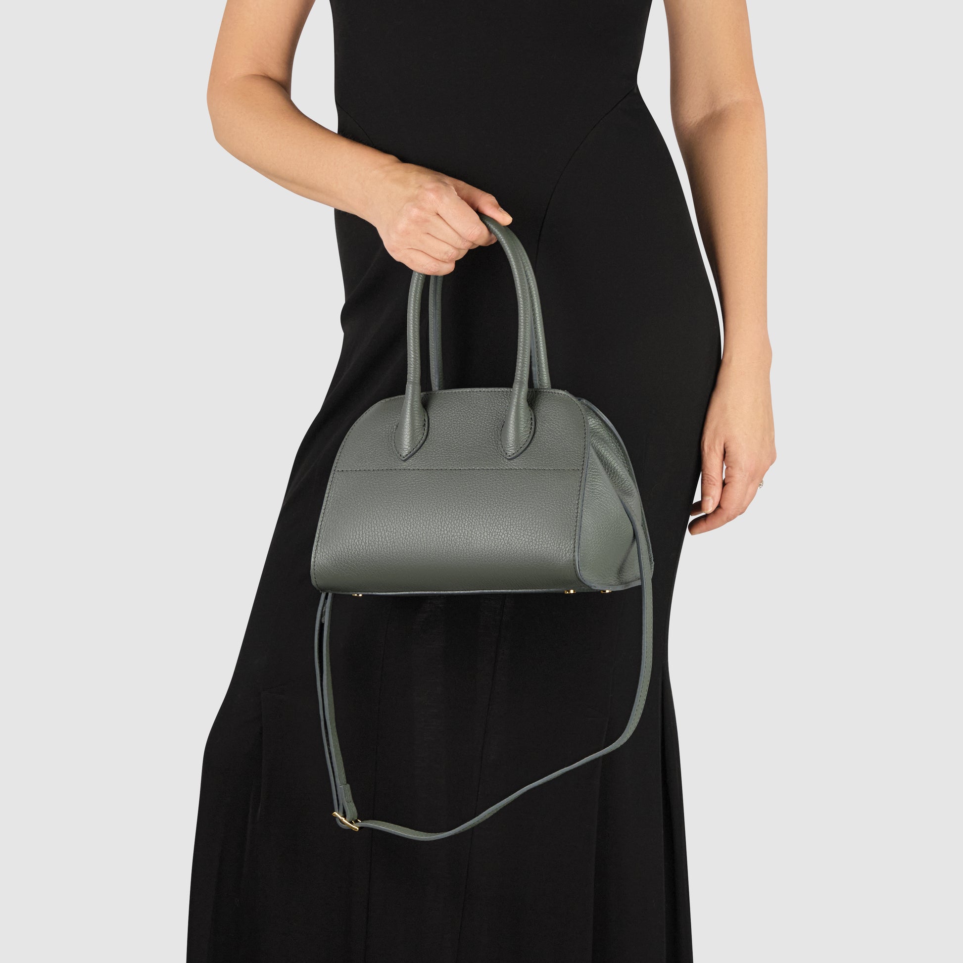 A person in a sleeveless black dress holds an Italian leather handbag with two handles and a shoulder strap against a plain light background.