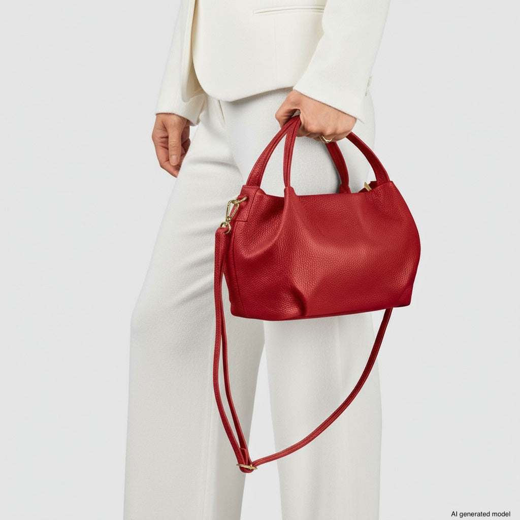 A person dressed in a white suit holds a dumpling-shaped, textured red handbag with a shoulder strap, standing against a plain light background.