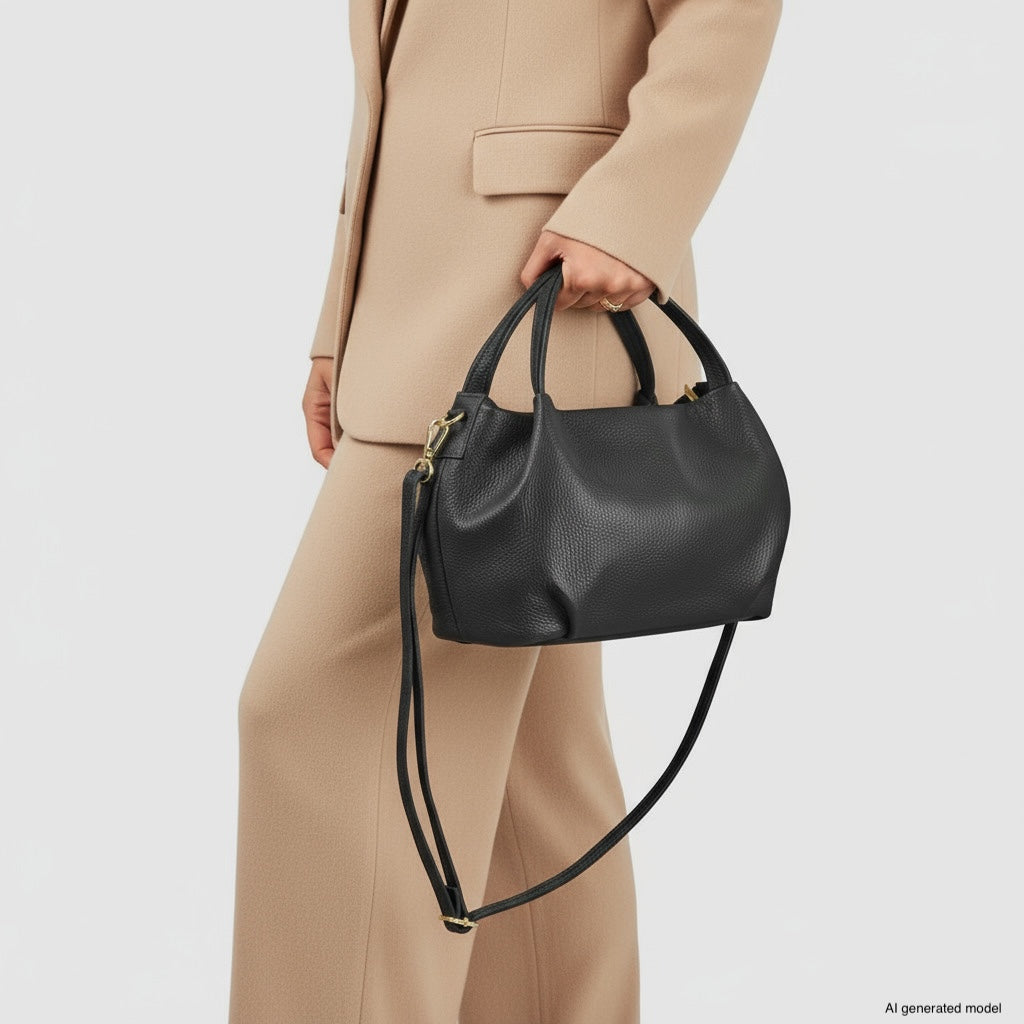 A person wearing a beige suit holds an Italian leather handbag with a shoulder strap. The dumpling-shaped bag stands out against the plain white background.