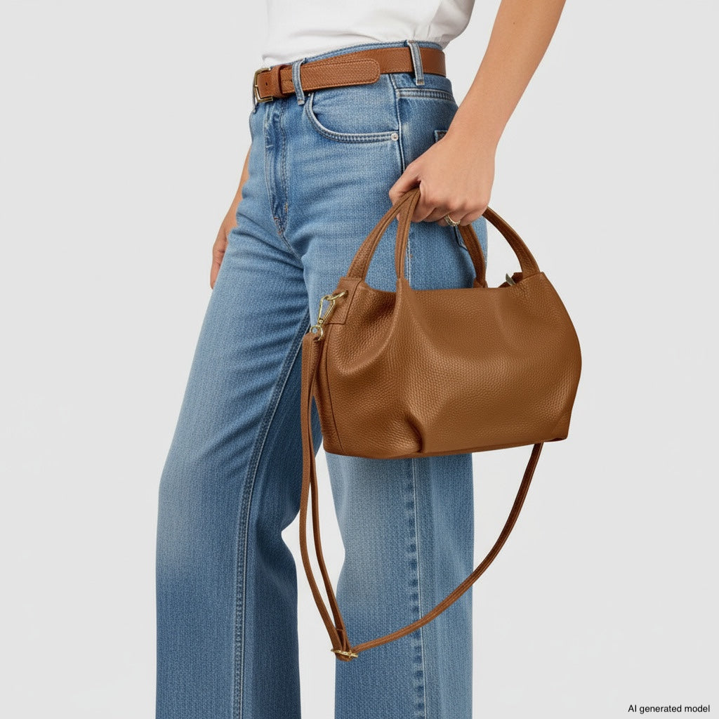 A person wearing high-waisted blue jeans and a brown belt holds an Italian leather handbag with a shoulder strap against a plain light background.